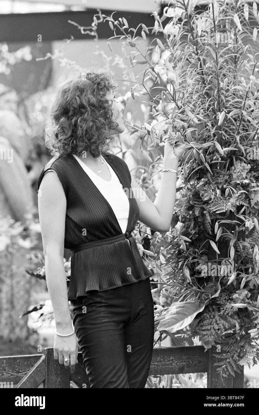 Ouverture Liliade Opening Openings Commissioning, Whizgle News, Dutch Desk, pays-Bas, 1950 - 2000 on 10-05-1979. Ce sont les éléments de l'image. La scène se déroule dans un jardin luxuriant et vibrant débordant de verdure. Une femme se tient au milieu d'un ensemble de plantes, son corps tourné légèrement sur le côté pendant qu'elle inspecte un arrangement de fleurs. Elle a des cheveux bouclés qui encadrent son visage doucement, ajoutant une touche d'élégance à son apparence. Sa tenue comporte un haut noir sans manches avec une texture plissée, qui contraste magnifiquement avec la chemise blanche nette en dessous. Cet ensemble est complemente Banque D'Images