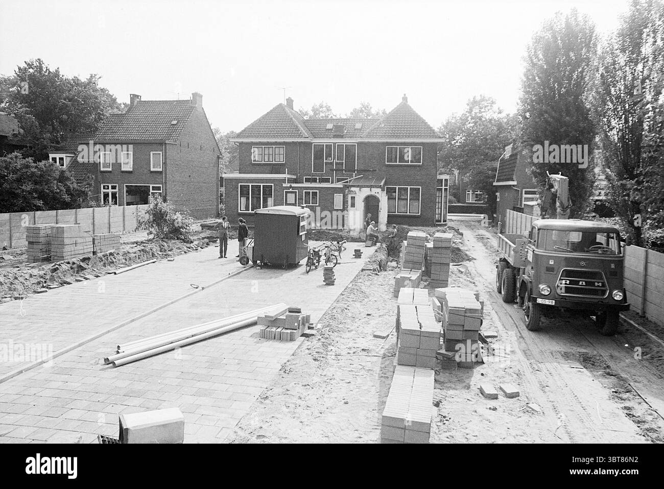 Road Makers, Whizgle News, Dutch Desk, pays-Bas, 1950 - 2000 sur 07-1971. L'image contient ces rubriques. La scène capture un chantier de construction dans une zone résidentielle. Au premier plan, des piles de briques rectangulaires soigneusement disposées bordent les deux côtés d'un chemin pavé qui semble mener à une grande maison de deux étages avec un toit incliné. La maison se caractérise par son architecture traditionnelle, avec de grandes fenêtres avec des cadres blancs et une porte d'entrée qui suggère une entrée accueillante. À gauche, quelques ouvriers de la construction sont engagés dans leurs tâches, posant éventuellement le bric Banque D'Images