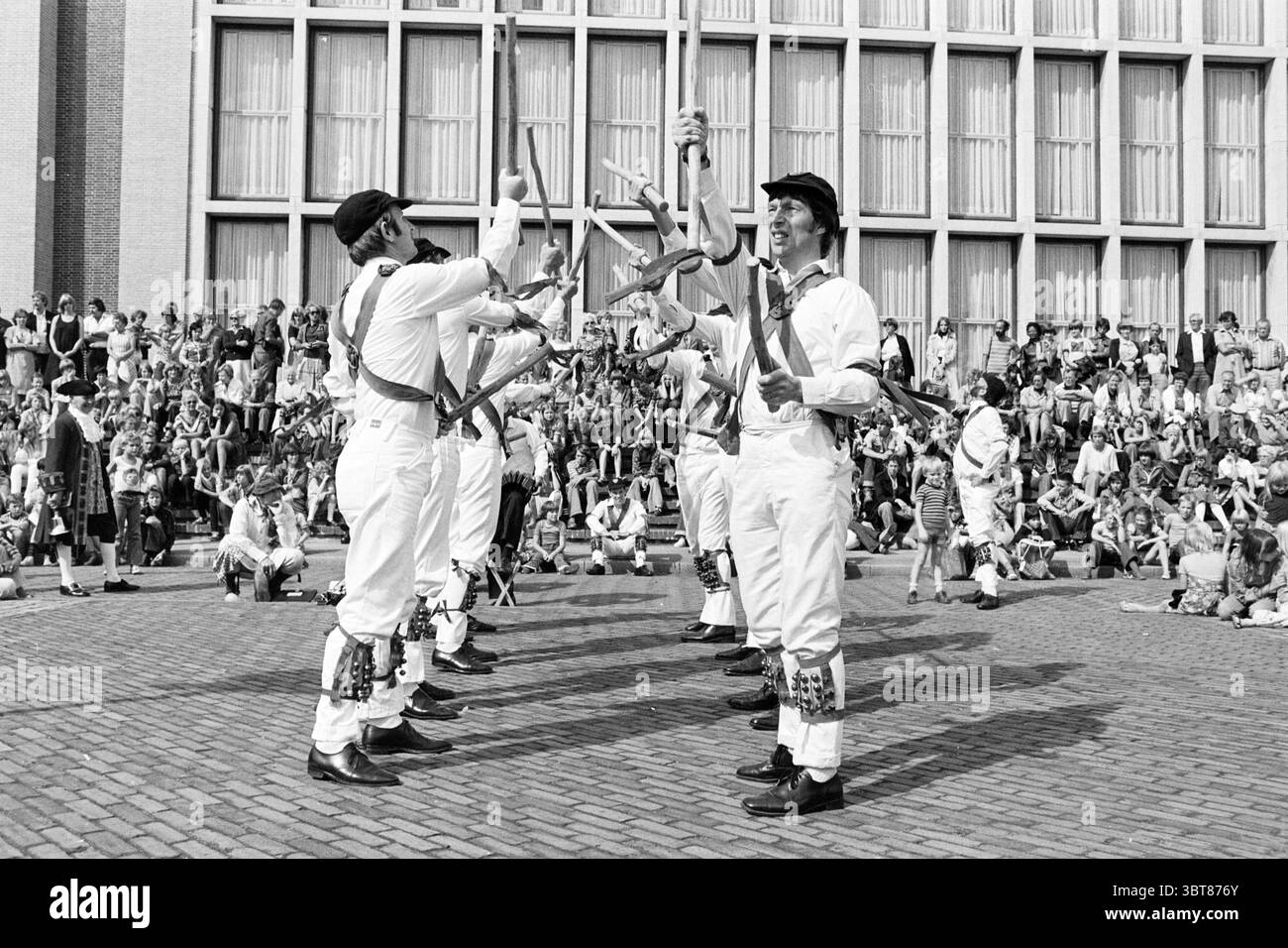 Morris danseurs à IJm. Danse, Whizgle News, Dutch Desk, pays-Bas, 1950 - 2000 le 03-09-1977. Ce sont les éléments de l'image. La scène capture un événement en plein air animé et plein d'activité. Un groupe d'hommes vêtus d'une tenue blanche traditionnelle se tient bien en évidence au premier plan, chacun portant des chapeaux noirs et équipé de ceintures colorées drapées sur leur corps. Ils sont engagés dans une performance synchronisée, jonglant habilement et lançant divers objets, probablement des bâtons ou des matraques, haut dans les airs. Les expressions sur leurs visages véhiculent un mélange de concentration et de joie, showcasin Banque D'Images