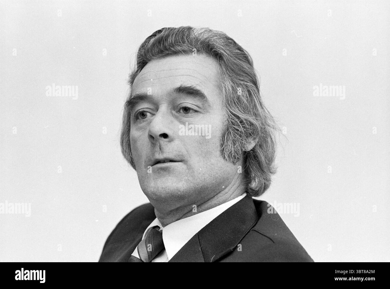 Rector Mr. Boers Sancta Maria College éducation personnelle, Whizgle News, Dutch Desk, pays-Bas, 1950 - 2000 le 06-12-1976. L'image inclut ces rubriques. Un homme avec une apparence distinguée est assis, représenté de profil. Ses cheveux, plus épais et légèrement ondulés, sont une nuance de gris clair, avec des pattes visibles encadrant son visage. Il porte un costume sombre, complété par une chemise de couleur claire et une cravate rayée, ce qui ajoute une touche de formalité à sa tenue. L'arrière-plan est une teinte douce et neutre qui met l'accent sur les traits de son visage. Son expression est sérieuse mais contemplative, sugg Banque D'Images