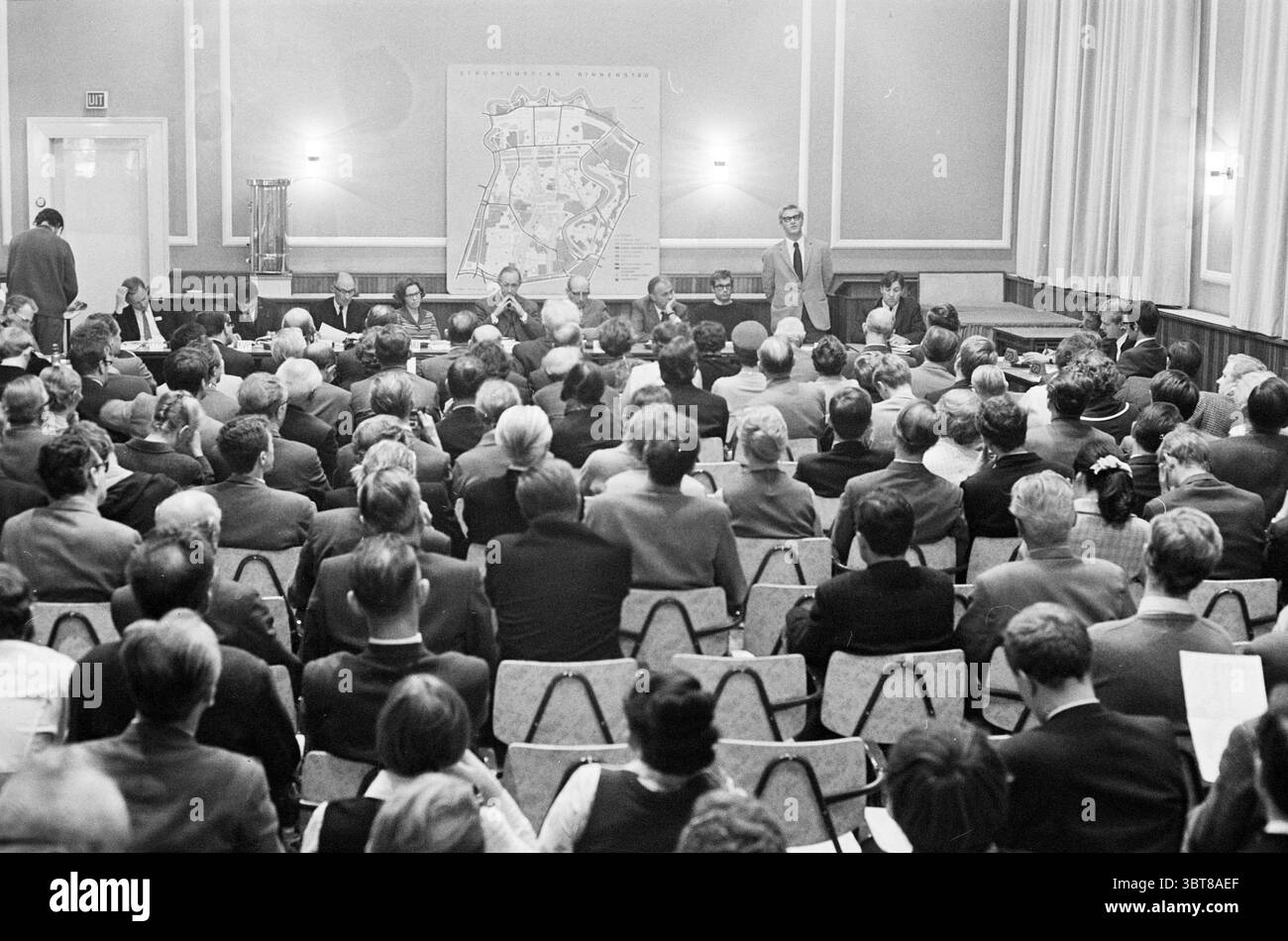 Teach-In City Center Meetings, Whizgle News, Dutch Desk, pays-Bas, 1950 - 2000 on 24-10-1969. L'image contient ces rubriques. La scène capture un grand rassemblement dans un cadre intérieur formel. Les rangées de chaises de couleur claire sont remplies d'un public diversifié, principalement des individus d'âge moyen et plus âgés, tous concentrés intentionnellement sur l'avant de la pièce. La foule est habillée dans un mélange de vêtements d'affaires, suggérant un événement professionnel ou communautaire. À l'avant, un groupe de personnes s'assoit derrière une longue table, engagé dans la discussion. Une personne se tient debout, s'adressant éventuellement au public, faisant des gestes Banque D'Images