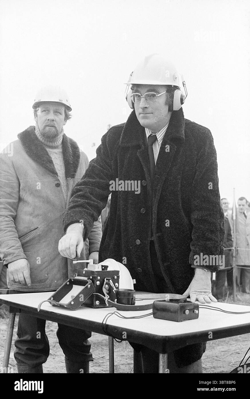 Maître sauteur et conseiller au premier coup de feu dans A'dam le premier bruit dans le port des paradis D'A'dam, Whizgle News, Dutch Desk, pays-Bas, 1950 - 2000 le 17-11-1976. Ces rubriques apparaissent dans l'image. La scène capture deux hommes dans un cadre industriel marqué par un sentiment d'anticipation. Le personnage principal est habillé d'un manteau sombre et flou sur une tenue formelle, ses mains posées sur un petit mécanisme situé sur une table. Il porte un casque de protection et des écouteurs, ce qui indique qu'il met l'accent sur la sécurité en dépit des dangers potentiels. Son expression est sérieuse, véhiculant peut-être de la concentration ou de la détermine Banque D'Images
