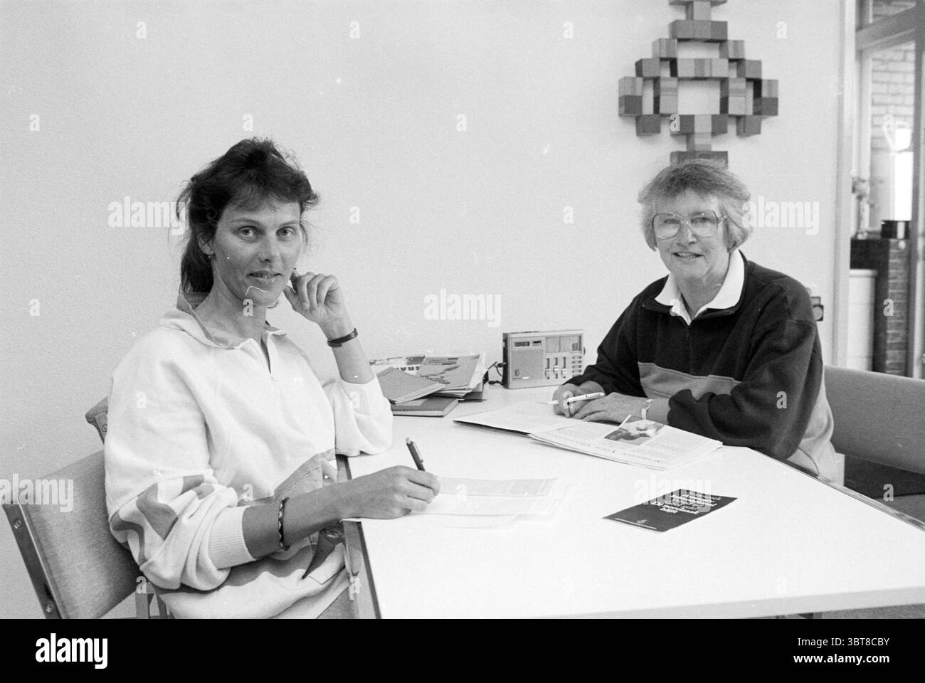 MRS Trip from Amnesty International Hillegom Hillegom, Whizgle News, Dutch Desk, pays-Bas, 1950 - 2000 on 22-05-1990. Ces rubriques apparaissent dans l'image. La scène se déroule dans un intérieur lumineux et minimaliste, caractérisé par un mur de couleur claire qui améliore l'ouverture de l'espace. Au premier plan, deux femmes sont assises à une simple table blanche. L'une d'elles a les cheveux foncés et ondulés et porte un sweat-shirt blanc décontracté ; elle tient un stylo et semble être engagée dans la pensée ou la conversation, reposant son menton sur sa main. L'autre femme a les cheveux courts et clairs et est habillée d'un darke Banque D'Images