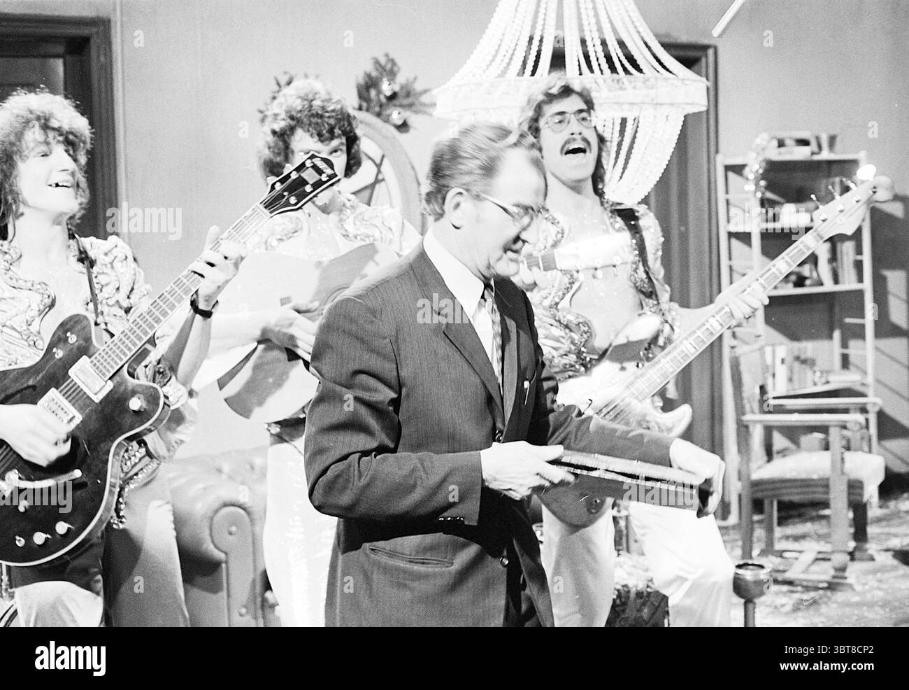 Groupe 'Circus' et Ir. E. van der Pik avec le disque 'No Beer but Buttermilk' à v. Oekels disco corner of the VPRO' Television, Whizgle News, Dutch Desk, pays-Bas, 1950 - 2000 le 27-12-1974. Ces rubriques sont illustrées dans l'image. La scène capture une performance musicale animée dans un cadre lumineux et de style rétro. Au premier plan, un homme vêtu d'un costume sombre et pointu tient un objet, peut-être un petit instrument ou un accessoire, avec une expression focalisée. Ses lunettes captent la lumière, ajoutant une qualité réfléchissante à son apparence. Derrière lui, un trio de musiciens respire l’enthousiasme et l’énergie. Ils Ar Banque D'Images