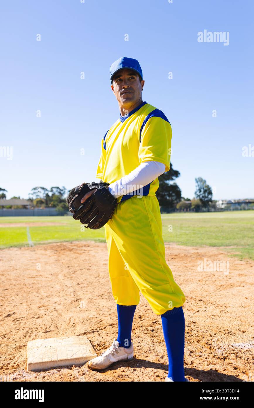 Athlète masculin debout sur le terrain près de la base, tenant un gant, portant un uniforme jaune et une casquette bleue Banque D'Images