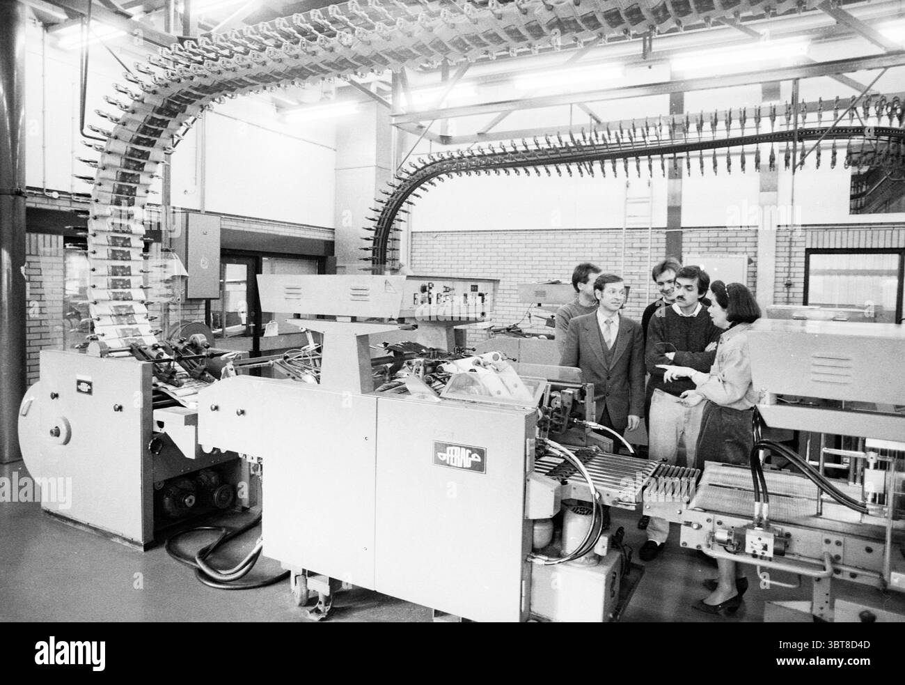 Deux Russes visitent l'imprimerie Haarlems Dagblad, Whizgle News, Dutch Desk, pays-Bas, 1950 - 2000 le 13-12-1990. L'image contient ces rubriques. La scène révèle un environnement industriel spacieux caractérisé par sa disposition expansive et son design fonctionnel. Au cœur de la composition se trouve une grande machine complexe qui comprend une gamme de composants mécaniques, y compris des bandes transporteuses et des pièces techniques complexes. La machine présente un éclat métallique, avec différentes nuances de gris et d'argent qui dominent la palette de couleurs. Il y a des éléments de bleu et de jaune visibles, éventuellement Banque D'Images