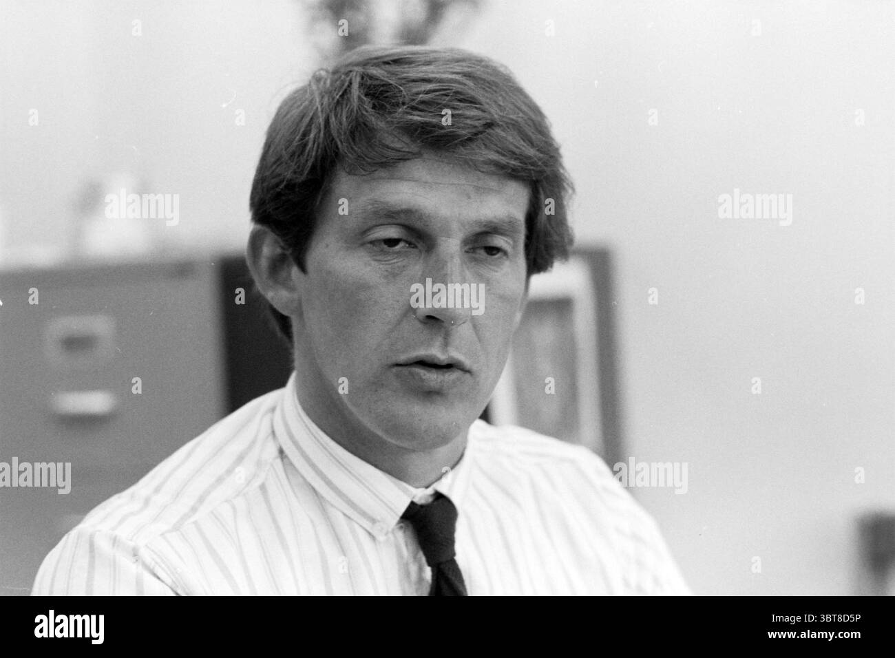 M. Populier Unie BLHP Schiphol Schiphol Schiphol, Whizgle News, Dutch Desk, pays-Bas, 1950 - 2000 on 12-06-1985. Ce sont les éléments de l'image. Dans un environnement de bureau légèrement éclairé, un homme est assis à un bureau, son expression contemplative et sérieuse. Il a des cheveux courts et touffus qui semblent légèrement désalignés, complétant son comportement concentré. Sa tenue est formelle mais simple : une chemise blanche rayée avec de fines lignes noires et une cravate sombre qui ajoute une touche de professionnalisme. L'arrière-plan révèle des indices d'un espace de travail organisé, avec des classeurs et des œuvres d'art encadrées ou des documents v Banque D'Images