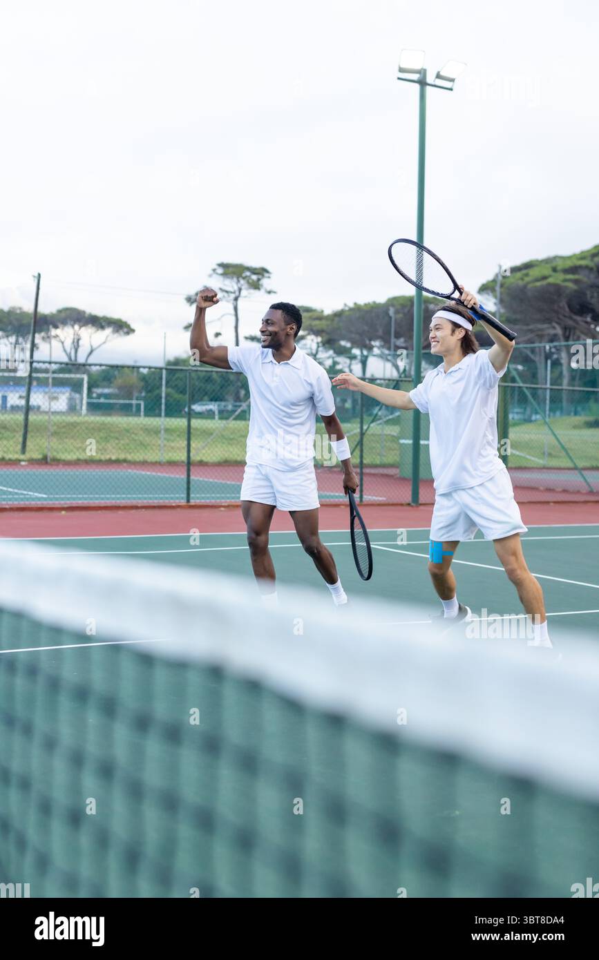 Divers amis masculins célébrant le point de victoire tout en marchant sur un court de tennis extérieur avec des raquettes Banque D'Images