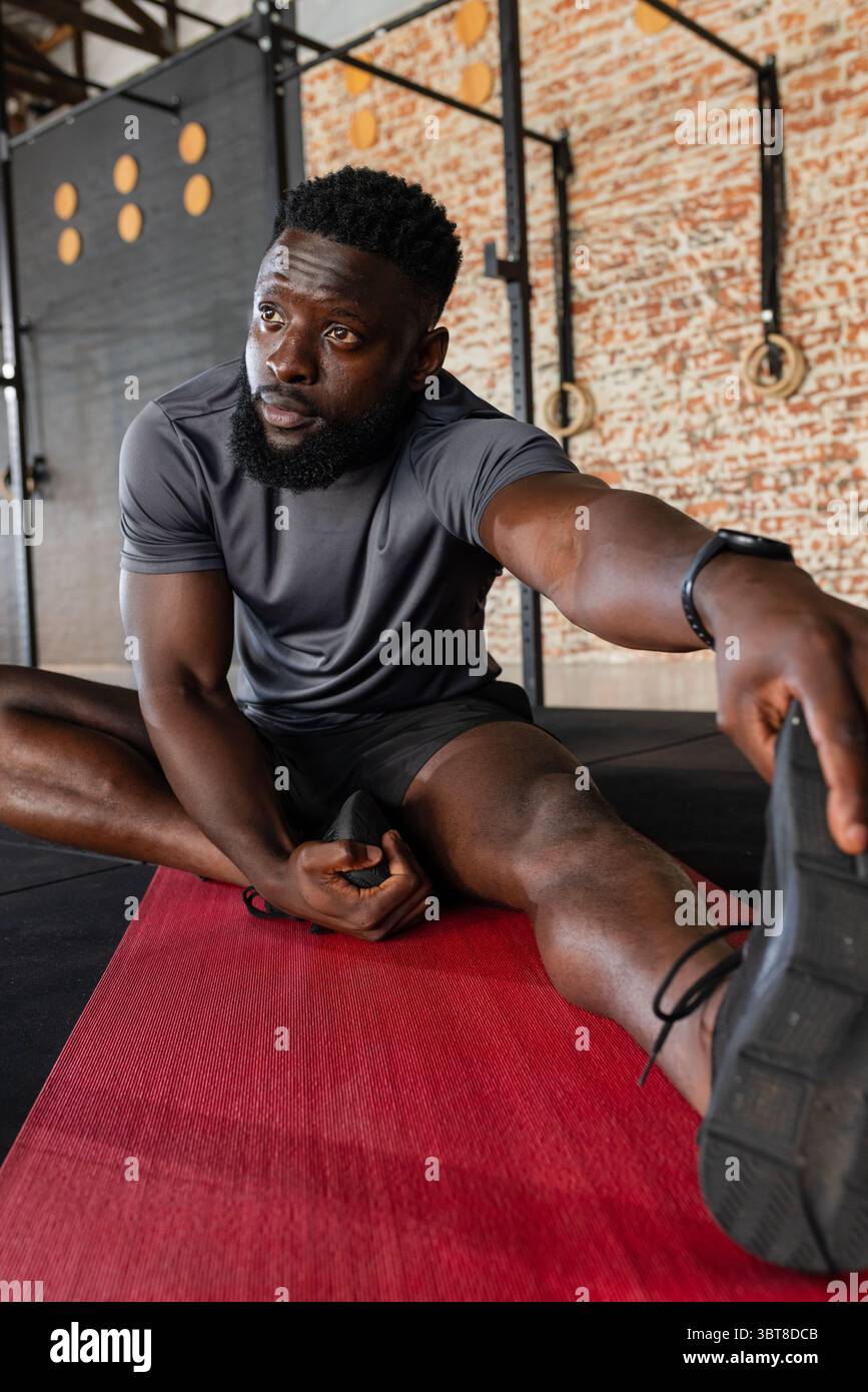 Homme afro-américain étirant la jambe sur le tapis rouge à la salle de gym tout en portant smartwatch et en utilisant des anneaux Banque D'Images