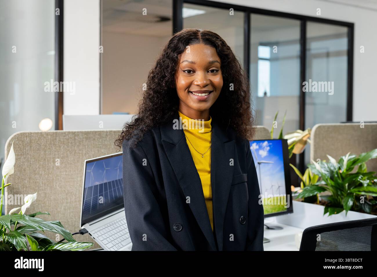 Femme afro-américaine debout et souriante dans un bureau ouvert au bureau avec des plantes de moniteur d'ordinateur portable Banque D'Images