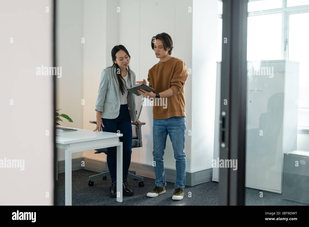Collègues chinois asiatiques s'appuyant sur le bureau blanc avec la plante en pot et examinant la tablette dans le bureau Banque D'Images