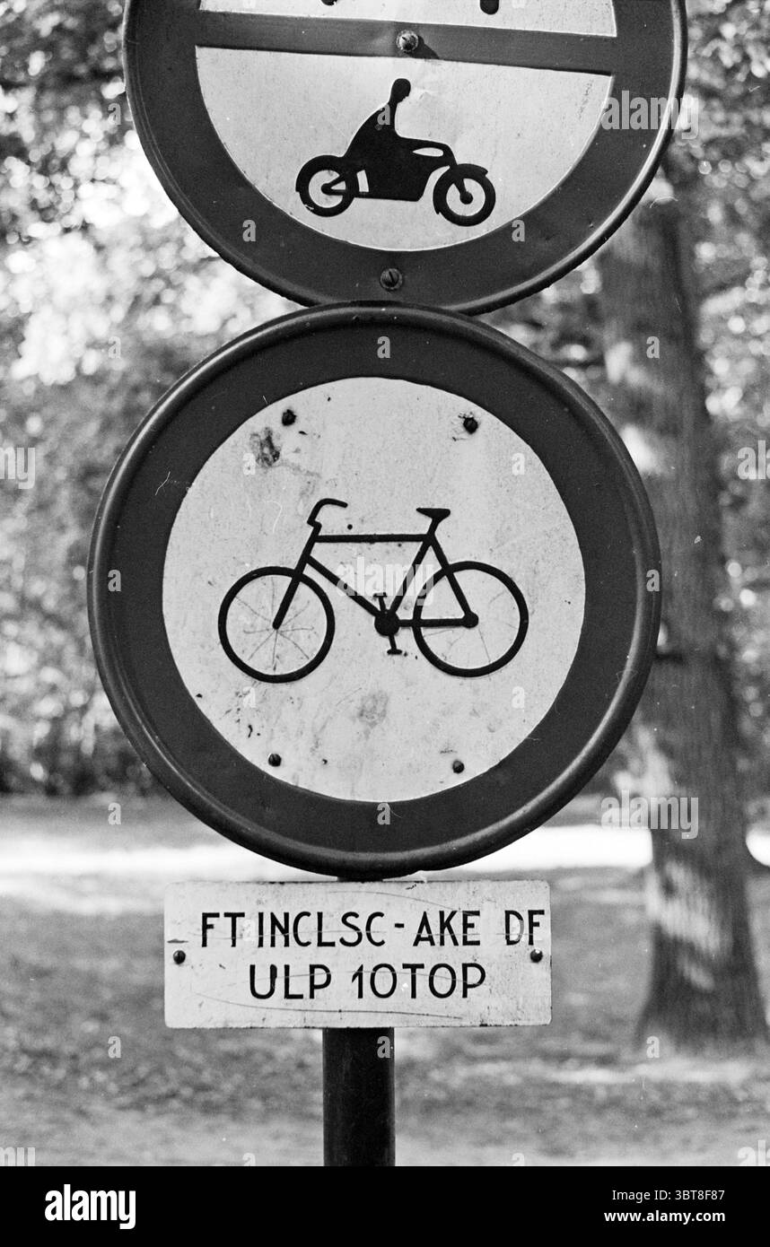 Panneau d'interdiction de bicyclette avec panneau inférieur : 'FT INCLUSC - AKE DF ULP 10TOP'., Whizgle News, Dutch Desk, pays-Bas, 1950 - 2000. Ces rubriques apparaissent dans l'image. Dans la scène, il y a deux panneaux circulaires montés sur un poteau. Le panneau supérieur représente un graphique d'une moto, rendu dans une forme simple et stylisée sur un fond blanc. En dessous, le deuxième panneau représente un vélo, également dans un style minimaliste, entouré d'une bordure circulaire. Le fond montre un cadre naturel, probablement un parc, avec des notes de verdure visibles, suggérant des arbres à proximité. Les couleurs des panneaux sont représentées par Banque D'Images