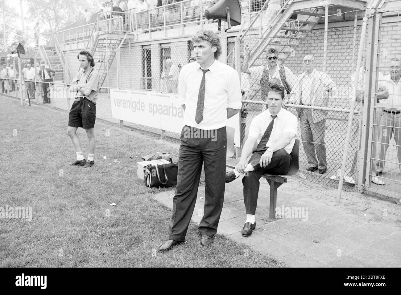 Match de football HFC Haarlem - P.S.V. match d'adieu de Soren Lerby qui a reçu un carton jaune et Guus Hiddink entraîneur du PSV. Voir NRC 07-05-1990 Haarlem Sportlaan pays-Bas, Whizgle News, Dutch Desk, pays-Bas, 1950 - 2000 le 06-05-1990. L'image montre ces rubriques. La scène capture un moment lors d'un événement sportif, probablement un match de football, qui se déroule dans un environnement extérieur ouvert. Au premier plan, deux hommes en tenue formelle se distinguent. L'un est positionné debout, les mains sur ses hanches, dégageant un sentiment d'autorité et de concentration, tandis que l'autre est assis, penché légèrement en arrière, paraissant plus détendu Banque D'Images
