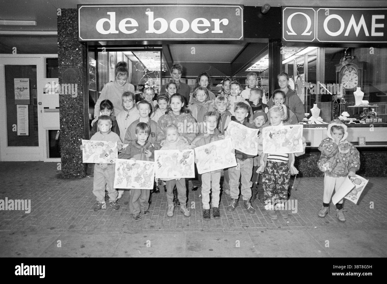Gagnants du concours de dessin par le bijoutier de Boer, Whizgle News, Dutch Desk, pays-Bas, 1950 - 2000 le 06-12-1991. L'image contient ces rubriques. Dans un cadre urbain animé, un groupe d'enfants se tient fièrement devant une devanture. Les enfants, un mélange diversifié d'âges et d'apparences, sont réunis en deux rangées, mettant en valeur leurs œuvres créatives. Chaque enfant porte des dessins aux couleurs vives, contrastant avec leurs vêtements discrets, qui vont de différentes nuances de brun et de gris à des teintes plus profondes. La scène est animée, avec les expressions des enfants montrant l'excitation et la fierté. Banque D'Images