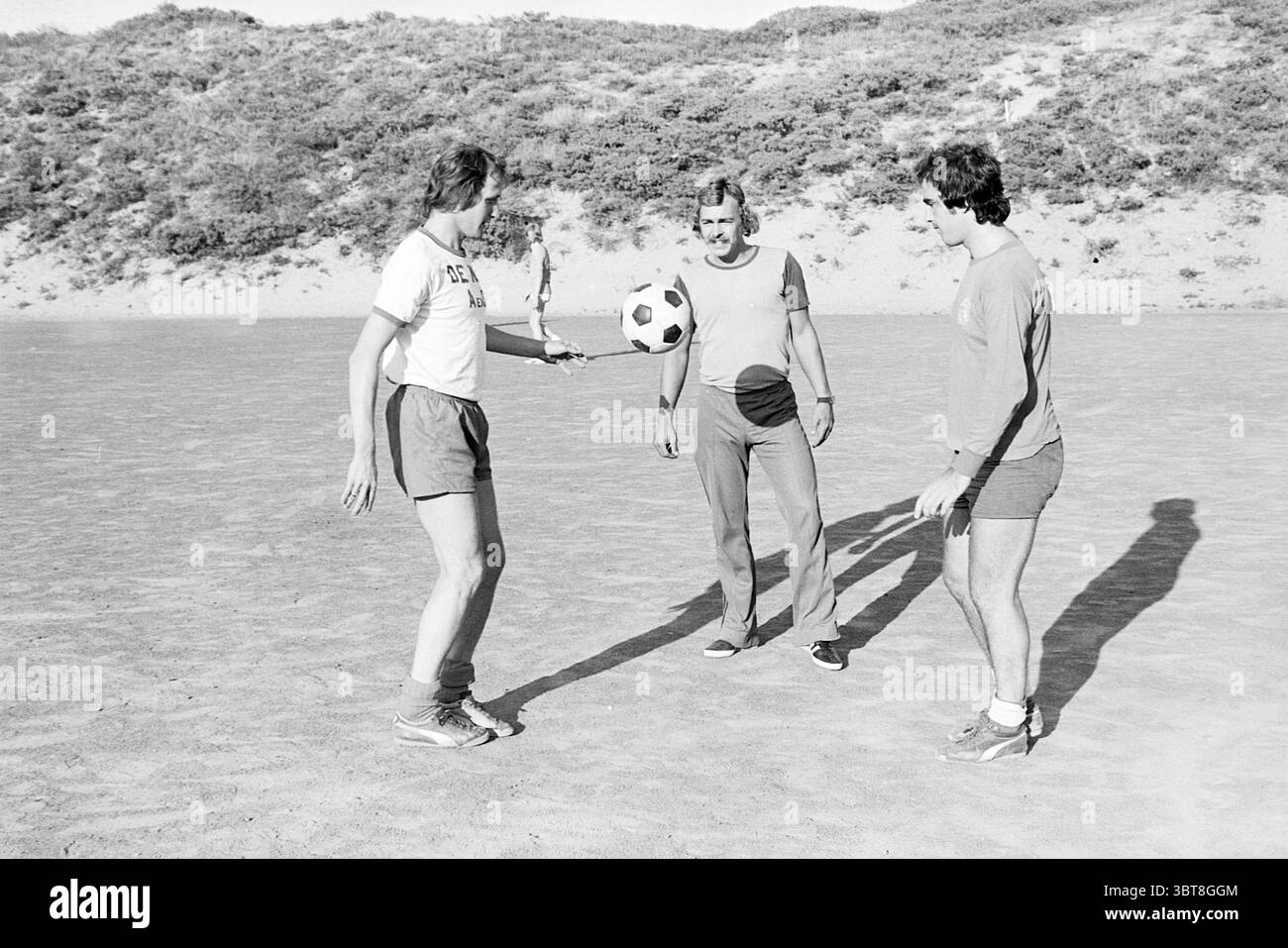 Les joueurs de football s'entraînent dans les dunes, Whizgle News, Dutch Desk, pays-Bas, 1950 - 2000 sur 07-1976. L'image montre ces rubriques. La scène se déroule sur un espace sablonneux et ouvert qui semble être un champ naturel ou une plage, marqué par de douces ondulations dans le sol. En arrière-plan, des collines couvertes de verdure luxuriante s'élèvent, leurs courbes projetant des ombres intéressantes à travers la région. Trois individus sont engagés dans une interaction ludique centrée autour d'un ballon de football. La première personne, une femme, se tient sur le côté gauche, portant un T-shirt de couleur claire et un short foncé, mettant en valeur son bui athlétique Banque D'Images
