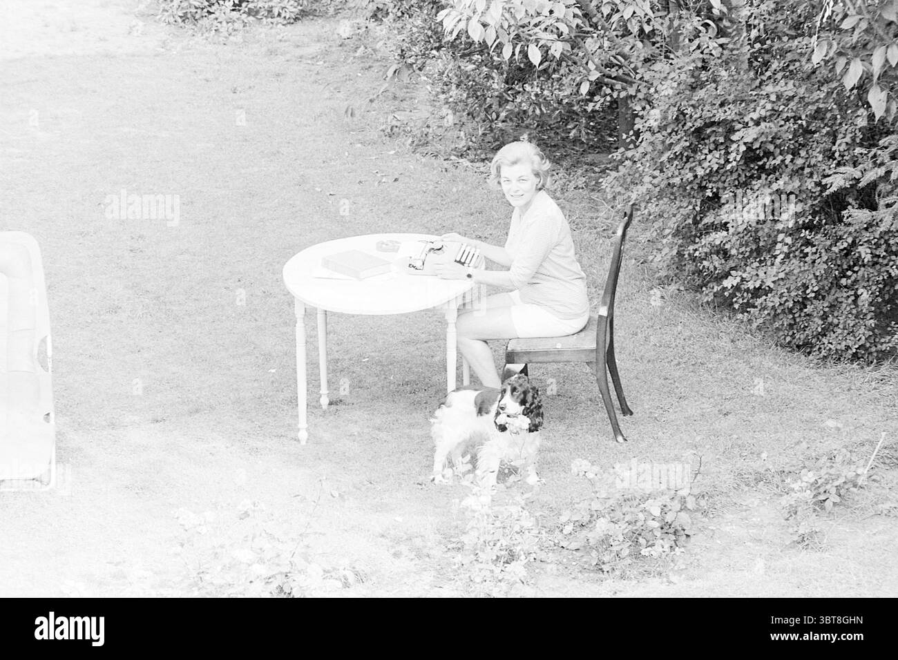MRS Dola de Jong écrivain d'Amérique en vacances ici personnes, Whizgle News, Dutch Desk, pays-Bas, 1950 - 2000 le 31-07-1963. L'image montre ces rubriques. La scène capture un cadre extérieur serein, baigné d'une lumière douce et diffuse qui crée une atmosphère douce et nostalgique. Une femme est assise à une petite table ronde, engagée dans une activité qui suggère concentration et créativité. Elle a un sourire chaleureux, ajoutant un sentiment de gaieté à l'instant. Elle est vêtue d'une tenue de couleur claire, qui se marie harmonieusement avec les teintes douces de l'environnement. Le tableau est simple, avec quelques objets Banque D'Images