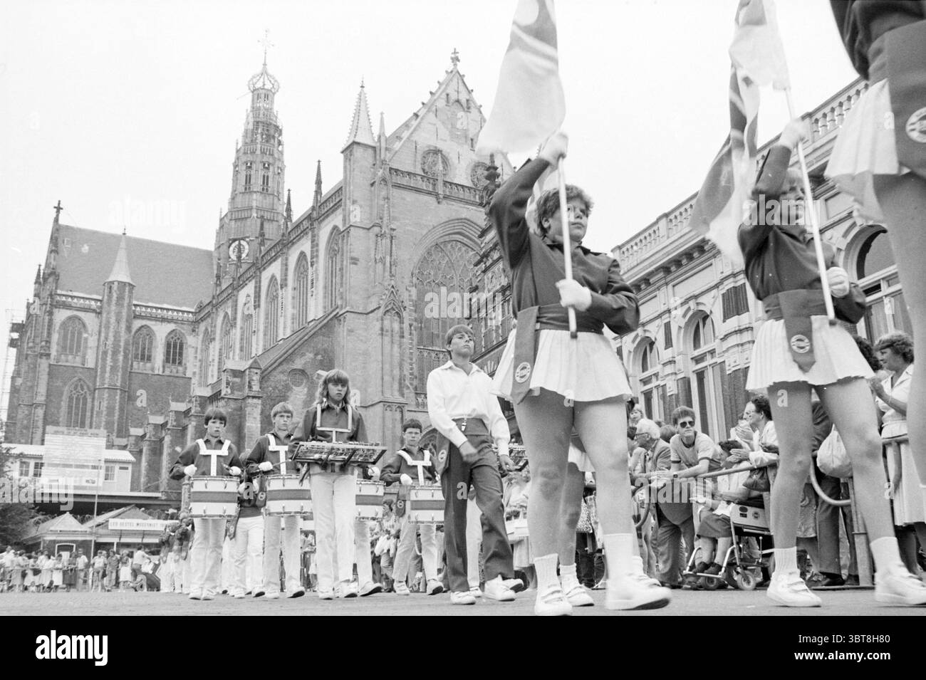 Tatouage Grote Markt Tattoo Haarlem Grote Markt pays-Bas, Whizgle News, Dutch Desk, pays-Bas, 1950 - 2000 le 22-06-1983. L'image contient ces rubriques. La scène présente une procession animée sur fond d'une église historique impressionnante caractérisée par ses hauts flèches et ses détails architecturaux complexes. La façade de l’église est ornée de grandes fenêtres, présentant des éléments gothiques qui élèvent la grandeur du cadre. Au premier plan, un groupe d'individus en uniforme, dont des enfants et des adolescents, défilent en formation. Ils portent des drapeaux de couleurs vives, EA Banque D'Images