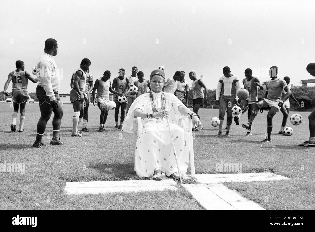 Coupe Haarlem : Medicine Man Nigeria and team Football, Whizgle News, Dutch Desk, pays-Bas, 1950 - 2000 le 30-07-1986. Ce sont les éléments de l'image. Sur un terrain d'athlétisme herbeux, une scène fortement composée se déroule, sur fond de ciel couvert qui fournissent une lumière douce et diffuse, créant une atmosphère silencieuse mais vibrante. À l'avant-garde, une figure royale vêtue d'une tenue blanche traditionnelle s'assoit en toute confiance sur une chaise. La tenue est agrémentée de motifs complexes, transmettant un sentiment d'importance culturelle et d'autorité. Sa posture est détendue mais commande, et elle porte Banque D'Images