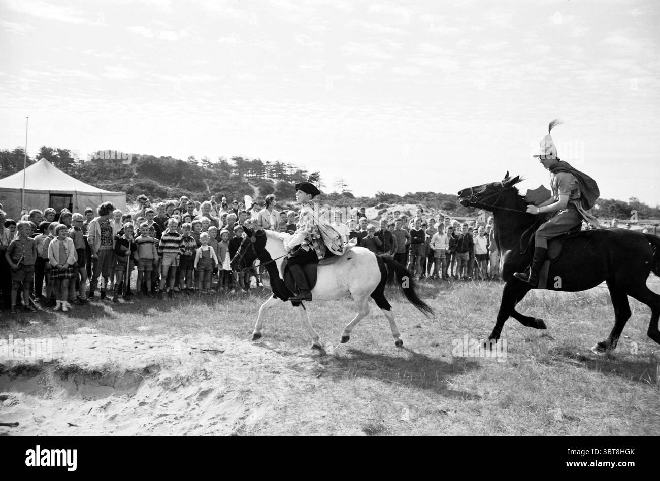 Ouverture par Ivanhoe Boys' Town IJmuiden Jeux de jeunes et de jeunes Jeux de vacances jours de vacances camp de vacances, Whizgle News, Dutch Desk, pays-Bas, 1950 - 2000 le 13-08-1962. Ces rubriques apparaissent dans l'image. Dans une scène animée en plein air, deux cavaliers à cheval galopent sur un terrain herbeux, attirant l'attention d'une foule nombreuse à proximité. Un cavalier est habillé dans une tenue de couleur claire, tandis que l'autre porte un uniforme plus formel orné d'un panache, suggérant un sens de l'autorité ou un but cérémoniel. Les chevaux sont distincts, l'un étant une bête vive de couleur claire et l'autre un hor sombre robuste Banque D'Images Ouverture par Ivanhoe Boys' Town IJmuiden Jeux de jeunes et de jeunes Jeux de vacances jours de vacances camp de vacances, Whizgle News, Dutch Desk, pays-Bas, 1950 - 2000 le 13-08-1962. Ces rubriques apparaissent dans l'image. Dans une scène animée en plein air, deux cavaliers à cheval galopent sur un terrain herbeux, attirant l'attention d'une foule nombreuse à proximité. Un cavalier est habillé dans une tenue de couleur claire, tandis que l'autre porte un uniforme plus formel orné d'un panache, suggérant un sens de l'autorité ou un but cérémoniel. Les chevaux sont distincts, l'un étant une bête vive de couleur claire et l'autre un hor sombre robuste Banque D'Images