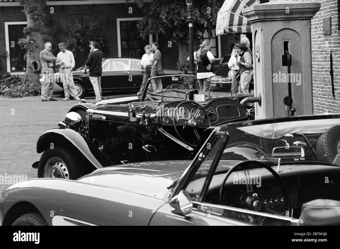 Vieilles voitures dues au rallye Oud Velsen, Whizgle News, Dutch Desk, pays-Bas, 1950 - 2000 le 08-05-1993. Voici les rubriques de l'image. La scène capture un charmant rassemblement en plein air, rappelant un spectacle de voitures anciennes ou un événement tranquille l'après-midi. Au premier plan, deux voitures vintage dominent la vue, leurs finitions brillantes et leurs designs élégants reflétant un sentiment de nostalgie. Une voiture, à la peinture noire profonde, révèle un tableau de bord poli avec des cadrans et des textures complexes, tandis que l'autre, dans une nuance de rouge vif, complète l'ambiance avec ses lignes sportives et épurées. Occupant le Banque D'Images