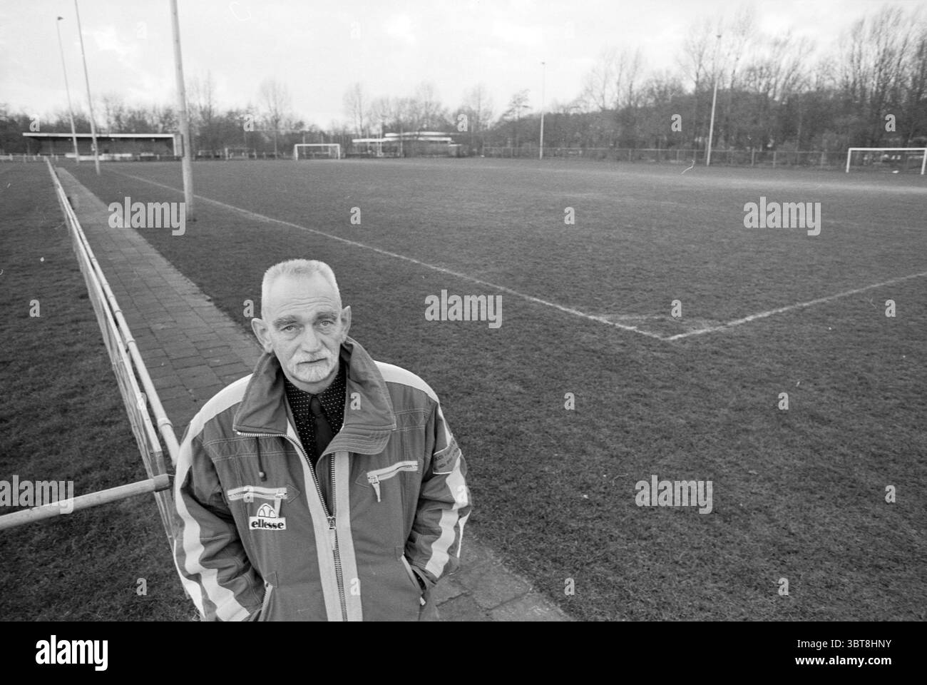 Mr. Votez pour Velsen, Whizgle News, Dutch Desk, pays-Bas, 1950 - 2000 le 16-02-1995. L'image inclut ces rubriques. Dans une scène monochrome, un homme âgé se tient bien en vue au premier plan, surveillant un vaste terrain de football vide. Il est habillé d'une veste décontractée zippée avec un logo, qui contraste avec les boutons sombres de sa chemise en dessous. Son expression est réfléchie, reflétant peut-être la nostalgie, alors qu'il regarde au loin. Le terrain de football s'étend derrière lui, marqué par une herbe fraîchement coupée et luxuriante qui a été bien entretenue, situé dans un grand espace ouvert. Deux buts a Banque D'Images