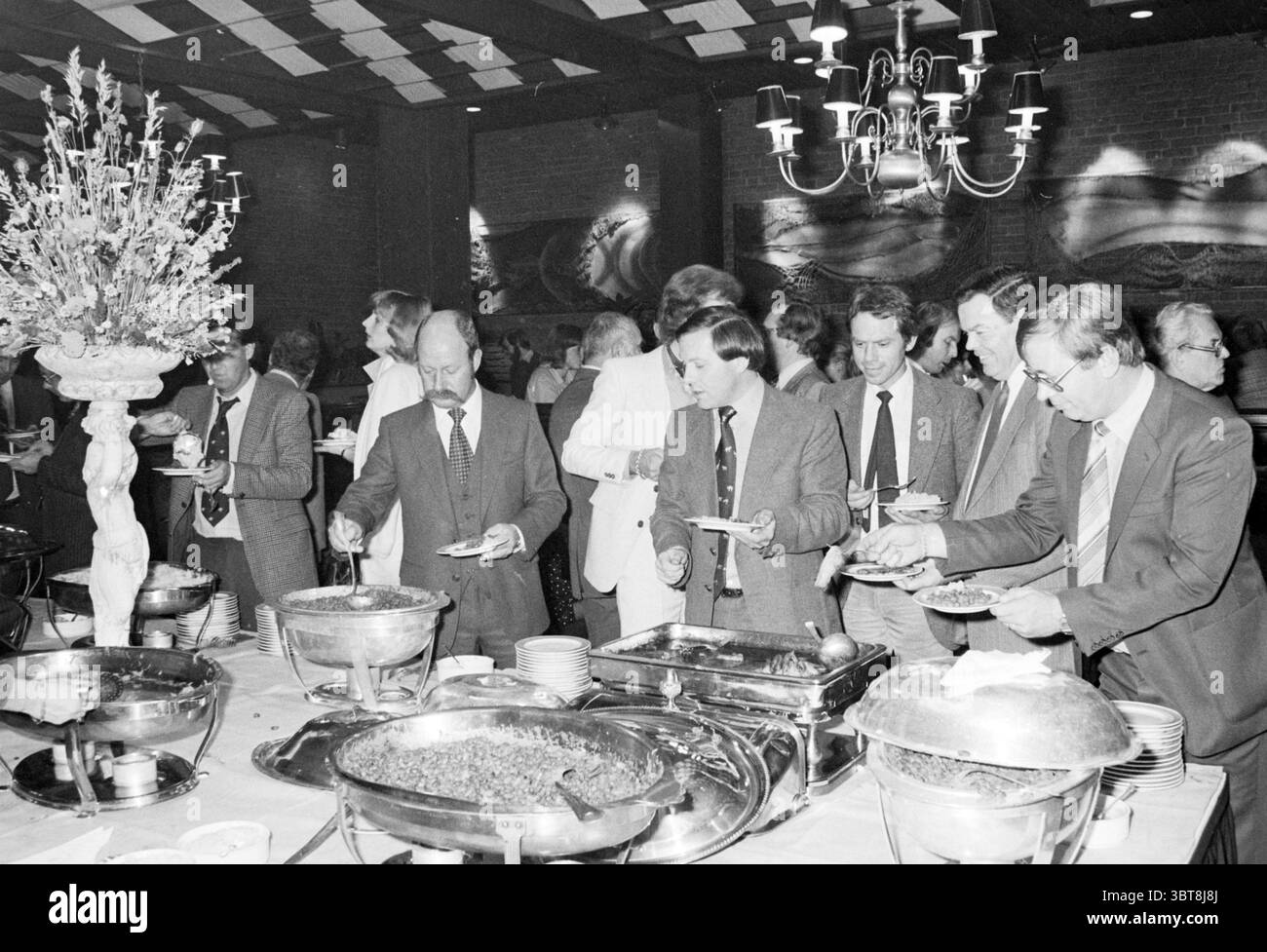 Admedia Fête d'automne à Schiphol Hilton Parties Schiphol, Whizgle News, Dutch Desk, pays-Bas, 1950 - 2000 le 28-10-1979. Ce sont les éléments de l'image. Une scène de banquet animée se déroule dans une salle chaleureusement éclairée, caractérisée par une ambiance élégante. Plusieurs hommes, habillés de costumes allant des tons foncés aux tons clairs, forment une ligne devant une vaste table de buffet. Les participants semblent engagés dans la conversation, montrant des expressions de plaisir et de camaraderie. Le buffet propose une variété de plats présentés dans des plateaux de service en métal brillant, reflétant la lumière ambiante. Les aliments incluaient Banque D'Images