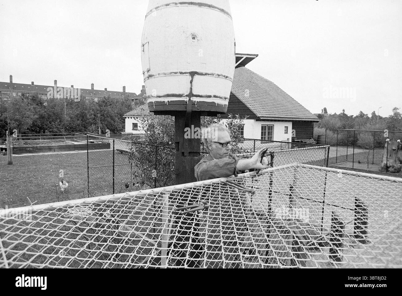 HR Van son chez animal Farm IJmuiden Persons, Whizgle News, Dutch Desk, pays-Bas, 1950 - 2000 on 03-10-1973. L'image contient ces rubriques. La scène capture un cadre extérieur tranquille avec un mélange d'éléments naturels et artificiels. Un homme se tient concentré près d'une grande structure cylindrique ressemblant à un réservoir d'eau ou une mangeoire à oiseaux. Il est engagé dans une activité, peut-être nourrir ou interagir avec les oiseaux. Sa tenue vestimentaire se compose de vêtements pratiques, adaptés au travail en plein air. Autour de lui se trouve une cour spacieuse, entourée d'une clôture en filet, ce qui permet la visibilité tout en gardant les oiseaux en sécurité. V Banque D'Images