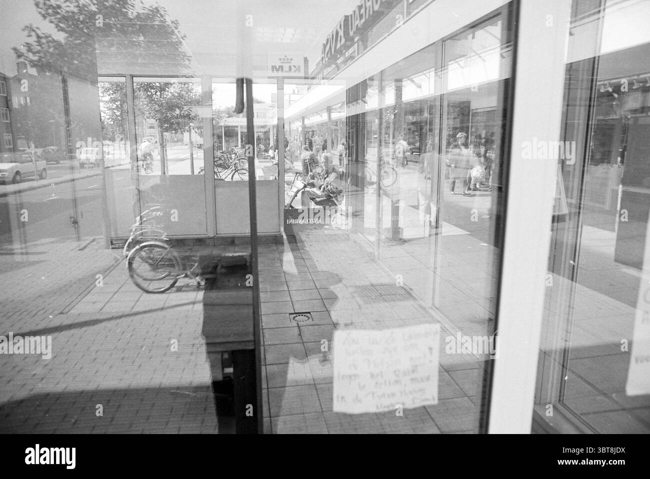 Magasin vide IJmuiden, Whizgle News, Dutch Desk, The Netherlands, 1950 - 2000 sur 23-08-1993. Voici les rubriques de l'image. La scène capture une vue de rue animée à travers le verre transparent, créant un effet visuel en couches. Au premier plan, les reflets des personnes et leurs mouvements sont perceptibles, indiquant une atmosphère animée. Des groupes d'individus se promènent devant, certains bavardant avec désinvolture tandis que d'autres semblent engagés dans leurs propres pensées. Le verre a un aspect légèrement trouble, réduisant la clarté mais ajoutant une qualité artistique aux reflets. En arrière-plan, les silhouettes de bi Banque D'Images