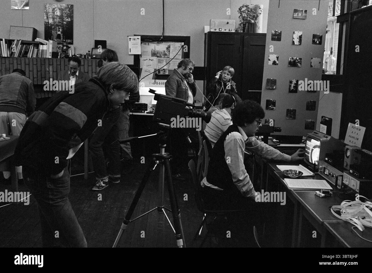Journée portes ouvertes radioamateurs Haarlem North Transmitters équipement de diffusion Haarlem pays-Bas, Whizgle News, Dutch Desk, pays-Bas, 1950 - 2000 on 29-10-1982. L'image inclut ces rubriques. La scène capture un cadre vivant et dynamique, rappelant un atelier ou un studio animé. Un groupe de personnes, hommes et femmes, participent activement à diverses activités, créant un sentiment de collaboration et de créativité. Au premier plan, un jeune homme se penche intensément vers une caméra posée sur un trépied. Ses cheveux courts et clairs et sa tenue décontractée suggèrent un look détendu mais focu Banque D'Images