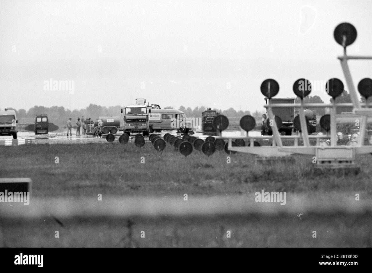 Bulderbaan Schiphol Schiphol, Whizgle News, Dutch Desk, pays-Bas, 1950 - 2000 on 24-06-1993. Ces rubriques apparaissent dans l'image. La scène capture une zone large et ouverte qui semble être un aérodrome ou une piste d'atterrissage. Le premier plan est dominé par une série de grands objets circulaires, probablement des marqueurs ou des signes, disposés en rangées nettes. Ces objets sont clairsemés et placés sur une toile de fond d'un sol mou et flou. Au milieu du terrain, plusieurs véhicules sont stationnés ou en mouvement. Parmi eux, quelques fourgonnettes se distinguent, suggérant un environnement de travail, tandis qu'un camion plus grand, appartenant peut-être à un service Banque D'Images