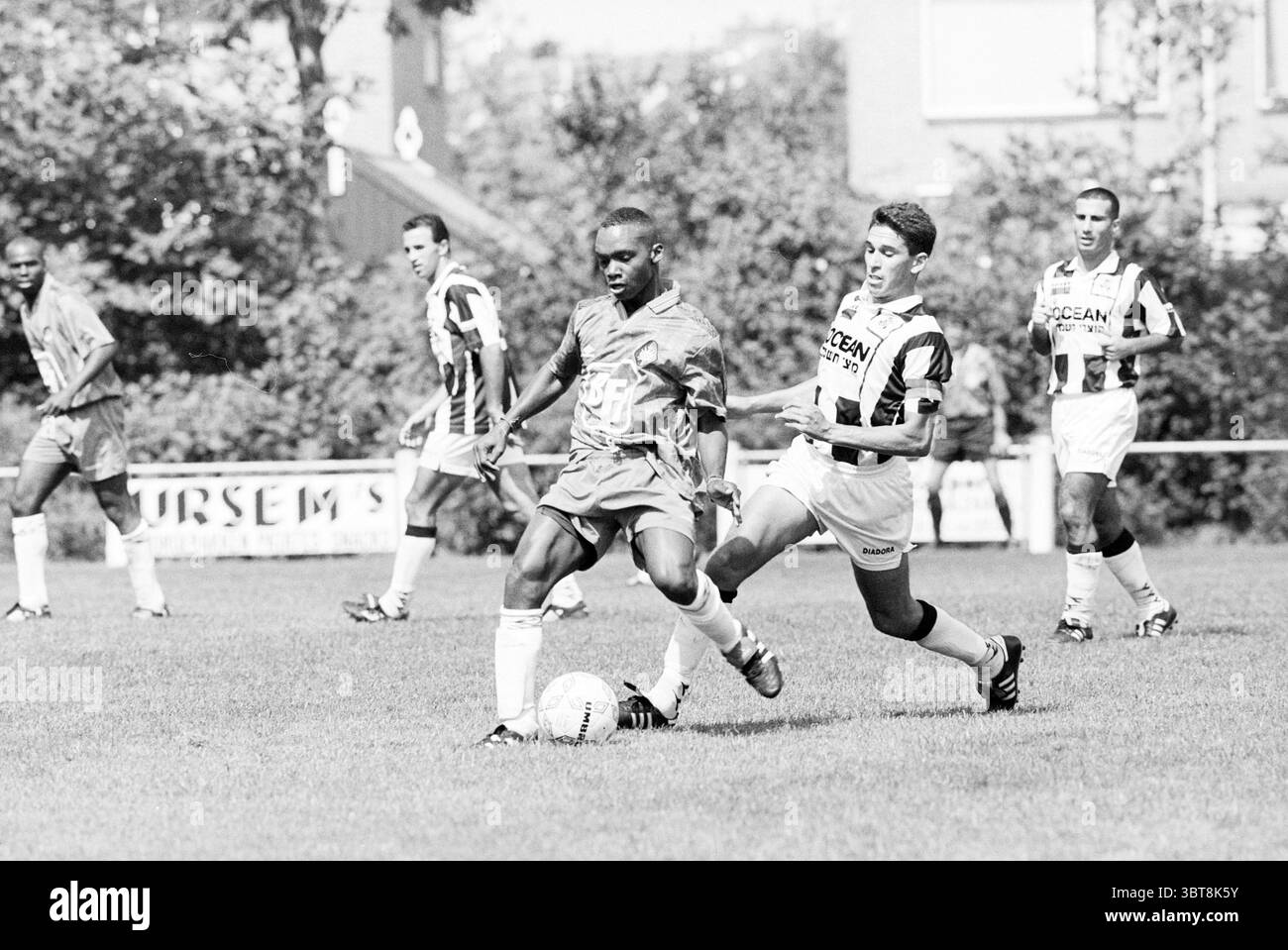 Football : Telstar - Israël, Whizgle News, Dutch Desk, pays-Bas, 1950 - 2000 le 18-07-1997. L'image contient ces rubriques. Dans un cadre extérieur dynamique, deux joueurs s'engagent dans un moment intense d'un match de football sur un terrain herbeux. La scène capture une interaction dynamique entre les athlètes, mettant l'accent sur leur athlétisme et leur concentration. Un joueur, vêtu d'un maillot sombre, dribble habilement le ballon, faisant preuve d'agilité lorsqu'il passe devant un adversaire. Cet adversaire, vêtu d'un uniforme rayé blanc et noir, tente de le défier, se positionnant stratégiquement pour faire un jeu. Le Banque D'Images