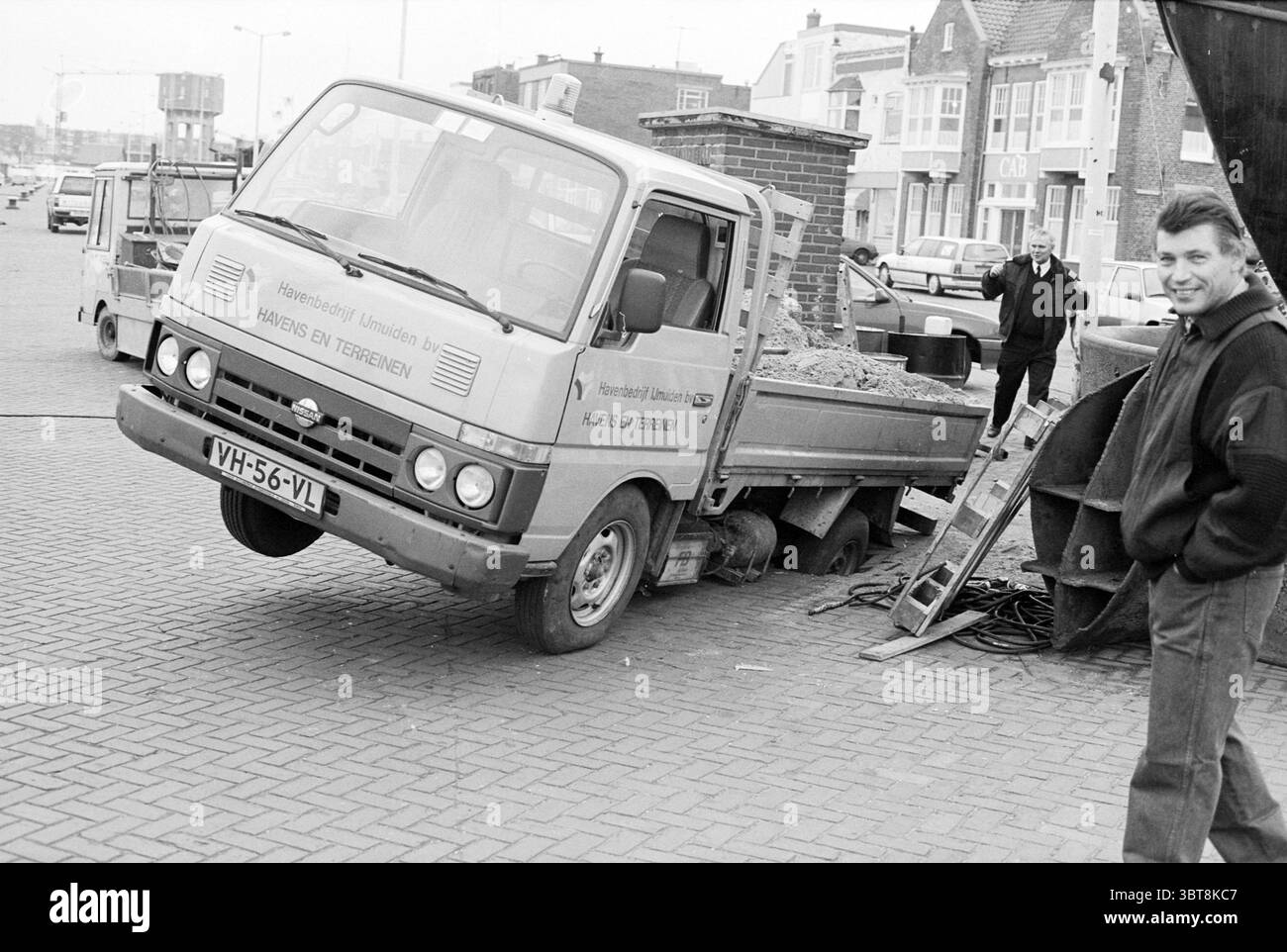 Voiture dans le trou IJm. IJmuiden pays-Bas, Whizgle News, Dutch Desk, pays-Bas, 1950 - 2000 le 31-01-1994. L'image inclut ces rubriques. La scène capture un moment dans une rue de la ville, caractérisée par une atmosphère un peu industrielle et décontractée. Un petit camion est bien visible, penché sur un côté, suggérant qu'il est en train de décharger ou de rencontrer une sorte de dysfonctionnement. Son dos est partiellement abaissé, avec une charge de gravier ou de boue qui se déverse, indiquant qu'il est engagé dans des travaux de construction ou d'entretien. Autour du camion, la zone pavée est marquée d'un dis Banque D'Images