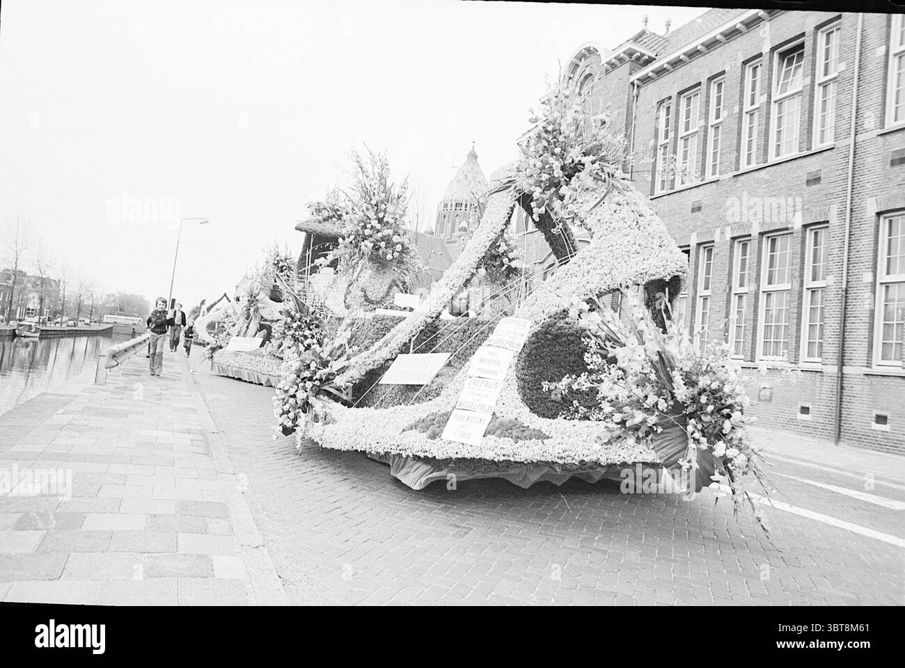 Défilé de fleurs Haarlem report défilé de fleurs avril 1972 Flower parade exposition de fleurs, Whizgle News, Dutch Desk, pays-Bas, 1950 - 2000 le 22-04-1972. Ces rubriques sont illustrées dans l'image. La scène présente un flotteur de parade florale élaboré, orné bien en évidence d'une variété de fleurs vibrantes, méticuleusement agencées pour former un design accrocheur. Le flotteur est grand et sinueux, serpentant son chemin le long d'un chemin pavé de briques à côté d'un canal. Les décorations florales comprennent un mélange de teintes vives, telles que des rouges audacieux, des violets profonds et des jaunes rayonnants, contrastant frappant avec le monochrome Banque D'Images