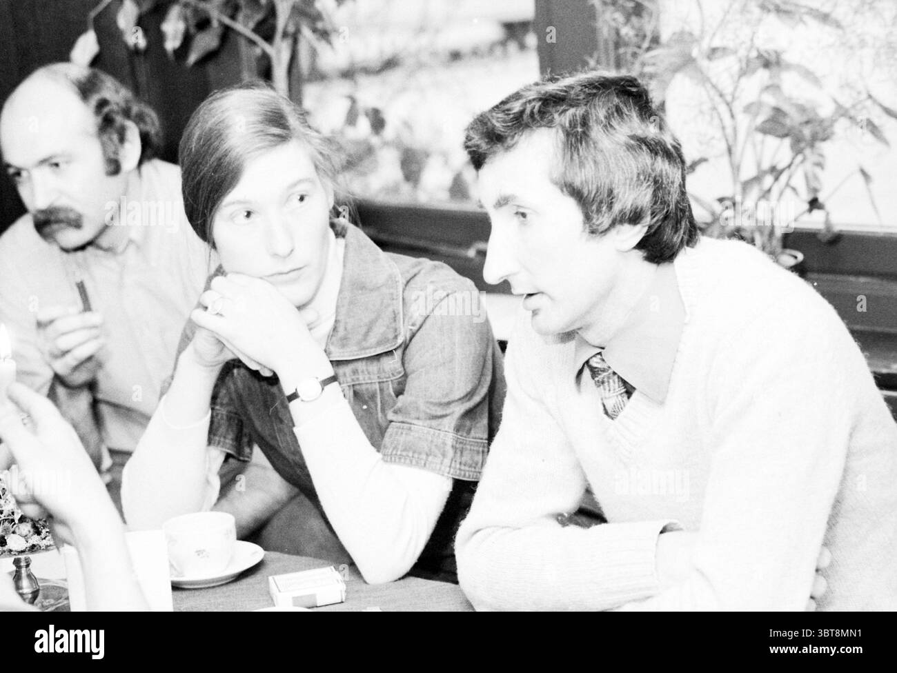 Diverses personnes en conversation, Whizgle News, Dutch Desk, pays-Bas, 1950 - 2000 le 07-05-1979. Ces rubriques apparaissent dans l'image. La scène se déroule dans un intérieur confortable, probablement un café ou un restaurant, caractérisé par des tons chauds et terreux. La lumière naturelle pénètre par les fenêtres voisines, créant une atmosphère douce et accueillante. Au premier plan, une jeune femme est assise engagée dans la conversation, son expression réfléchie alors qu'elle regarde vers un homme à côté d'elle. Elle a les cheveux longs et droits, et porte une robe en denim qui ajoute une touche décontractée mais élégante à son apparence. L'homme à côté d'elle, légèrement tourné, a Banque D'Images