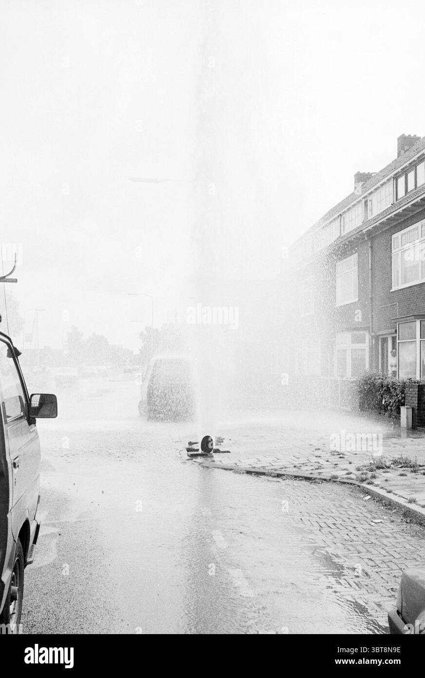 Fuite de tuyau d'eau Spaarndamseweg Haarlem Spaarndamseweg pays-Bas, Whizgle News, Dutch Desk, pays-Bas, 1950 - 2000 on 14-06-1990. L'image contient ces rubriques. La scène représente une rue dans un quartier résidentiel, caractérisée par un contraste saisissant entre les tons monochromes des environs et l'éclaboussure d'eau animée sortant d'un tuyau éclaté. Un puissant jet d'eau jaillit haut dans l'air, créant un voile brumeux qui domine la vue et baigne dans une lumière douce et diffuse. De chaque côté de la rue, il y a de modestes maisons de deux étages, dont les façades sont ornées Banque D'Images