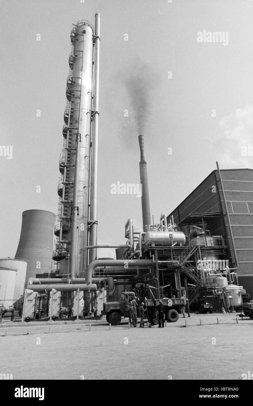 Nitric Acid Factory 5 Hoogovens, Whizgle News, Dutch Desk, pays-Bas, 1950 - 2000 le 23-04-1987. Voici les rubriques de l'image. La scène présente un paysage industriel caractérisé par une grande usine ou usine de transformation. Au-dessus des structures environnantes se dresse une colonne de distillation métallique haute, de forme cylindrique, reflétant le soleil dans des tons d'argent et de gris. À côté de lui se trouve une cheminée proéminente émettant un panache de fumée sombre qui contraste fortement avec le ciel clair, suggérant des opérations en cours au sein de l'installation. Le premier plan comprend un groupe de quatre individus Banque D'Images