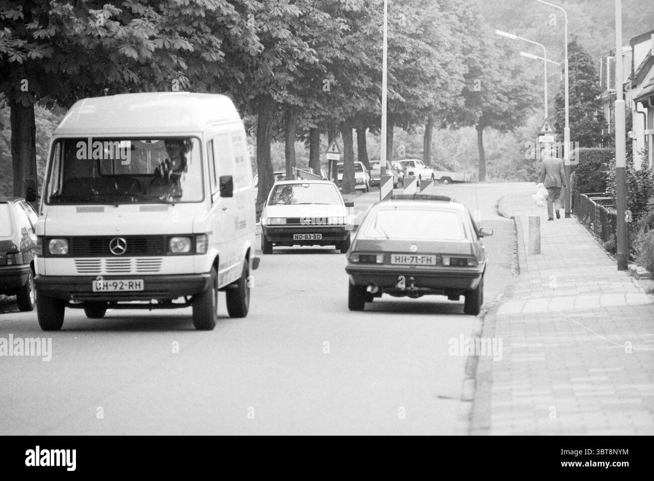Cycane Leidsevaart Vogelenzang. Routes construction de routes plantation de routes Vogelenzang Leidsevaart, Whizgle News, Dutch Desk, pays-Bas, 1950 - 2000 le 05-07-1988. L'image inclut ces rubriques. La scène représente une rue calme bordée d'arbres de chaque côté, leurs feuilles d'un vert doux, bien que le ton général soit silencieux, suggérant une journée nuageuse. Un fourgon de livraison blanc est placé bien en évidence sur le côté gauche, avec son avant légèrement incliné vers le spectateur, indiquant le mouvement. L'emblème Mercedes est visible, se démarquant de la peinture blanche du véhicule. Sur le côté droit du Banque D'Images