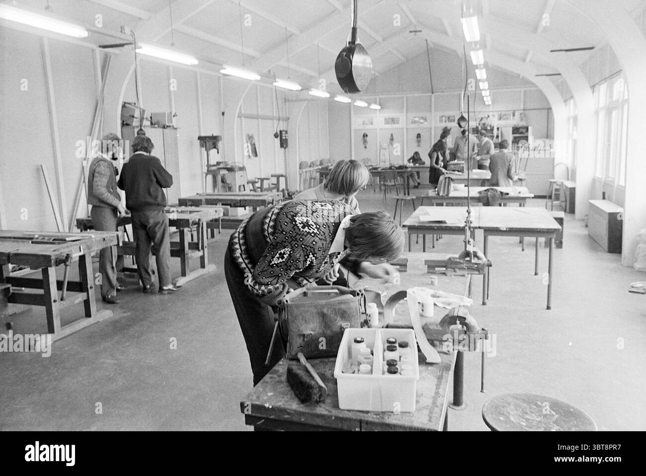 International Salle d'artisanat Ped. Academy Schools, Whizgle News, Dutch Desk, pays-Bas, 1950 - 2000 le 01-04-1971. L'image contient ces rubriques. La scène se déroule dans un atelier spacieux, caractérisé par son haut plafond voûté et sa lumière naturelle brillante qui traverse de grandes fenêtres. Les murs sont peints d'un blanc doux, tandis que le revêtement de sol est un béton gris pratique, faisant allusion à l'environnement industrieux. Au premier plan, une personne se penche sur un établi en bois, profondément concentré sur sa tâche. Leurs mains sont occupées à manipuler des outils et des matériaux, mettant l'accent sur la concentration. Cet individu est Banque D'Images