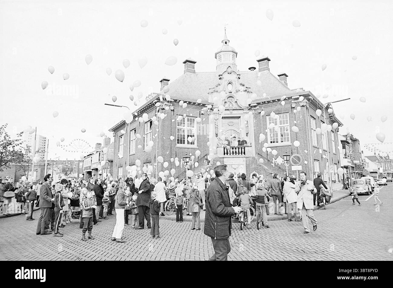 Zandvoort Town Hall Release Balloons., Whizgle News, Dutch Desk, pays-Bas, 1950 - 2000. Voici les rubriques de l'image. La scène capture un rassemblement animé dans un cadre urbain, caractérisé par une grande foule de personnes regroupées autour d'un bâtiment important. Cette structure, avec ses caractéristiques architecturales distinctives, a une façade charmante accentuée par des dessins ornés, lui donnant une sensation historique. Autour du bâtiment, une multitude d'individus couvre un spectre d'âges, tous engagés dans des interactions joyeuses. Beaucoup portent des vêtements décontractés, présentant divers motifs et col Banque D'Images