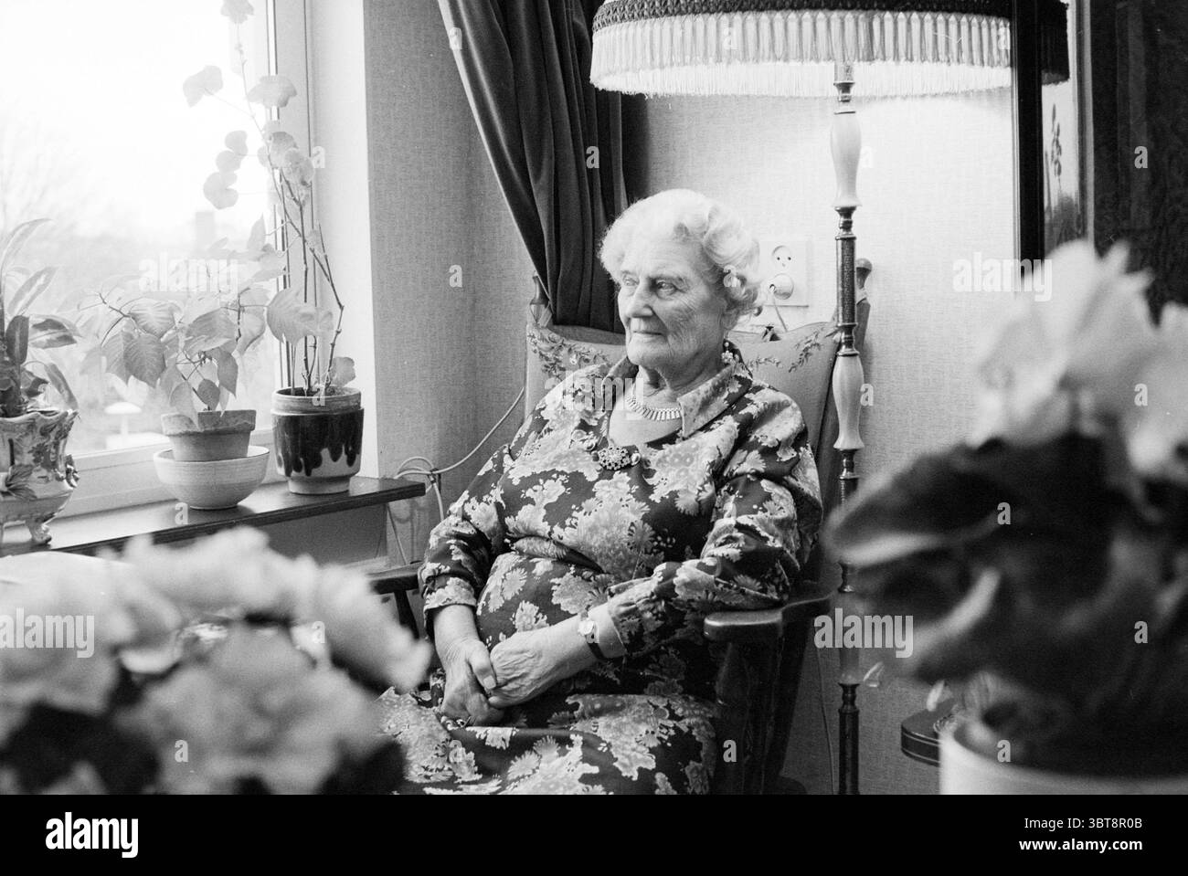 Portrait de femme 80+ assise dans le salon, Whizgle News, Dutch Desk, pays-Bas, 1950 - 2000 sur 08-12-1989. Ce sont les éléments de l'image. Dans un cadre intérieur confortable, une femme âgée s'assoit gracieusement dans un fauteuil en bois, dégageant une présence calme. Elle est habillée d'une robe à motifs floraux qui met en valeur des tons doux de rose et de bleu, s'harmonisant avec l'ambiance chaleureuse et accueillante de la pièce. Ses cheveux sont coiffés dans des boucles douces, et elle a une expression douce et sereine sur son visage, suggérant sagesse et expérience. Autour d'elle sont des plantes vertes luxuriantes, avec quelques fleurs en pot qui ajoutent spl Banque D'Images