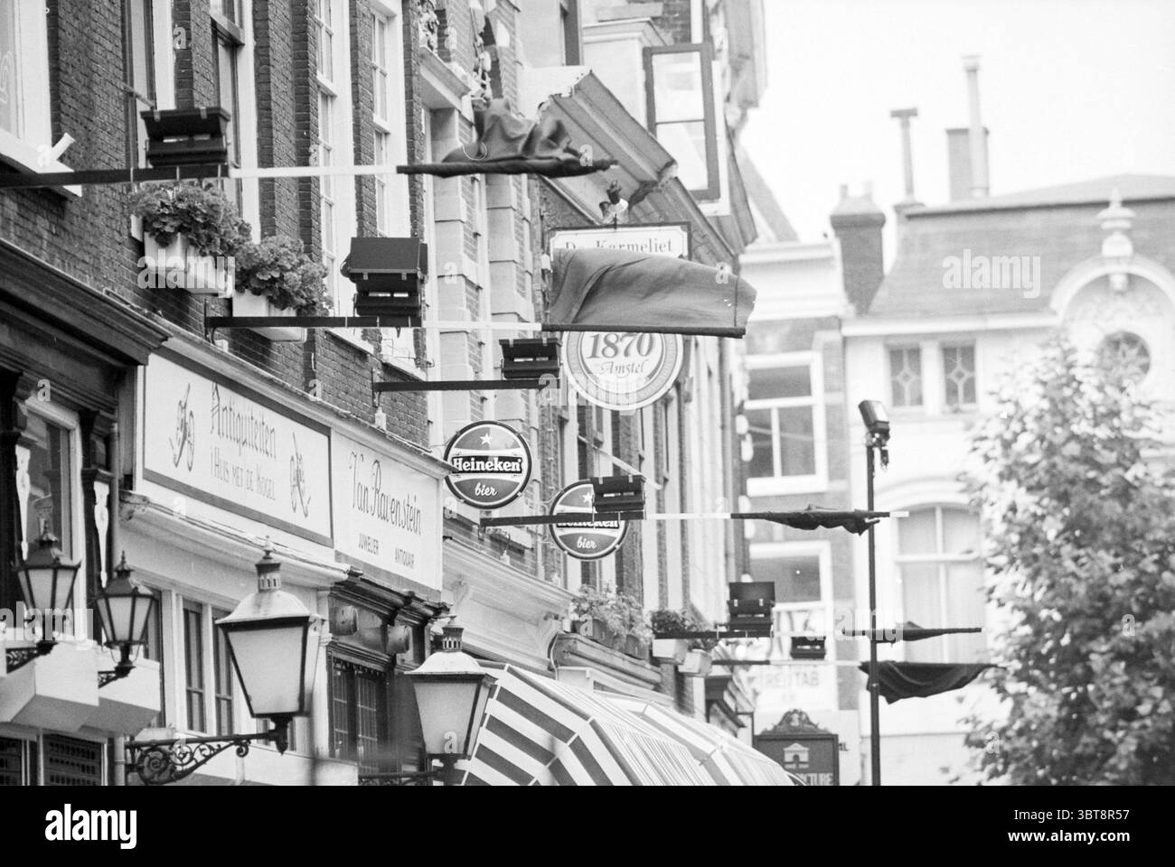 Drapeaux noirs concernant le départ et la démolition de Firma Enschedé Haarlem Spekstraat pays-Bas, Whizgle News, Dutch Desk, pays-Bas, 1950 - 2000 le 09-09-1993. L'image inclut ces rubriques. La scène présente une rue urbaine animée bordée de bâtiments présentant un mélange de styles architecturaux, mettant en valeur un charme historique. Les façades sont ornées d'une variété de signes et de décorations, faisant allusion aux diverses entreprises logées à l'intérieur. Plusieurs panneaux suspendus sont proéminents, certains circulaires et d'autres rectangulaires, présentant diverses couleurs et dessins, avec un accent notable sur de Banque D'Images