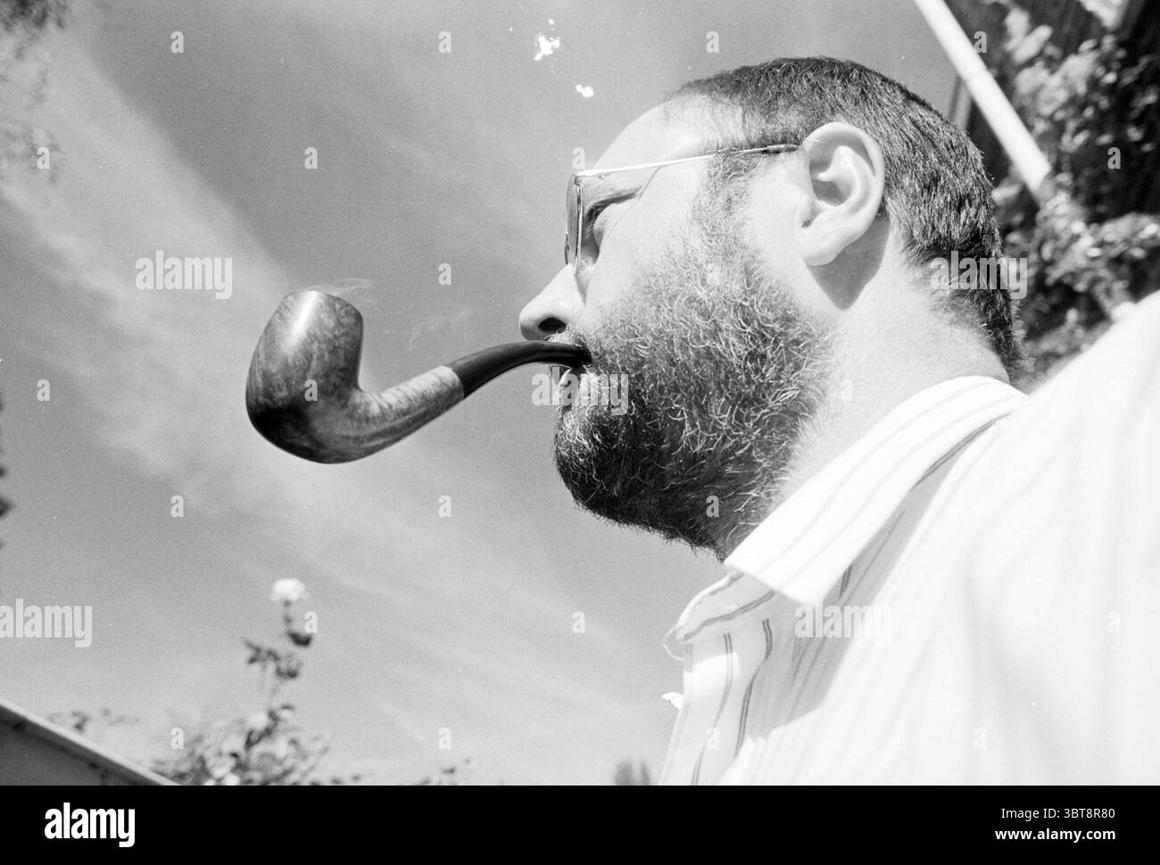 Henk van Dorp Pipe Smoking World Championships, Whizgle News, Dutch Desk, pays-Bas, 1950 - 2000 le 17-08-1993. L'image contient ces rubriques. La scène capture le profil latéral d'un homme à la barbe robuste, partiellement éclairé par la lumière naturelle du soleil. Sa tête est légèrement inclinée vers le haut, et une pipe à fumer classique repose entre ses lèvres, émettant un léger brin de fumée qui se recourbe élégamment dans l'air. L'homme porte des lunettes de soleil rondes, ajoutant une touche de sophistication à son apparence. En arrière-plan, des contours doux et flous de verdure peuvent être discernés, faisant peut-être allusion à un jardin ou un parc Banque D'Images