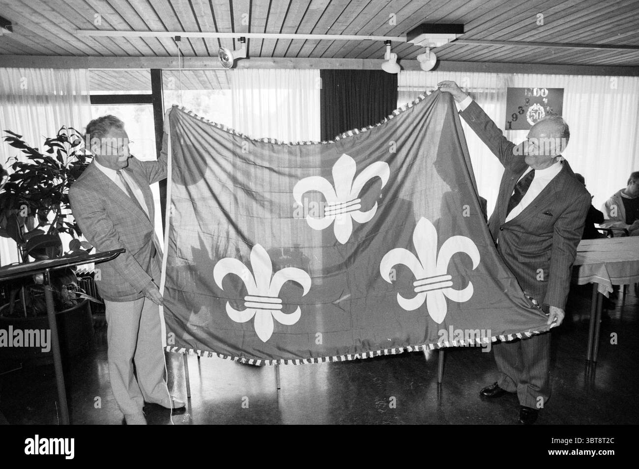 City flag 5 ans Theatre club Beverwijk remise et transferts Beverwijk pays-Bas, Whizgle News, Dutch Desk, pays-Bas, 1950 - 2000 le 25-09-1988. L'image contient ces rubriques. Deux individus sont au centre de la scène, chacun portant un drapeau élaboré avec un dessin proéminent avec trois symboles de fleur de lis disposés dans un motif symétrique. Le drapeau est vibrant, affichant des couleurs rouges et dorées profondes, qui contrastent avec les tons neutres de leur tenue et de l'environnement. Les deux hommes se tiennent côte à côte avec des expressions de fierté et de camaraderie. L'un est habillé Banque D'Images