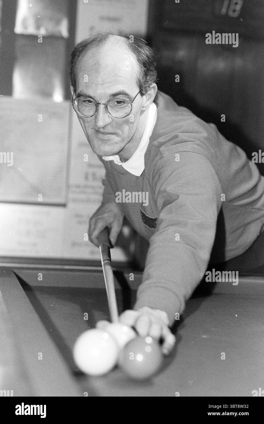 Joueur de billard, Whizgle News, Dutch Desk, pays-Bas, 1950 - 2000 sur 01-1988. Ces rubriques apparaissent dans l'image. La scène met en scène un individu concentré dans un décor de billard. La personne, un homme avec une récolte amincie de cheveux et de lunettes, est habillée d'un pull de couleur foncée au-dessus d'une chemise à col. Son expression reflète la concentration comme il se penche légèrement en avant, soulignant son intention sur le jeu. Devant lui se trouve une table de billard en feutre vert, avec deux boules pleines, une blanche positionnée vers le bord tandis qu'une autre boule blanche repose légèrement en avant. Le bâton de queue, saisi firml Banque D'Images