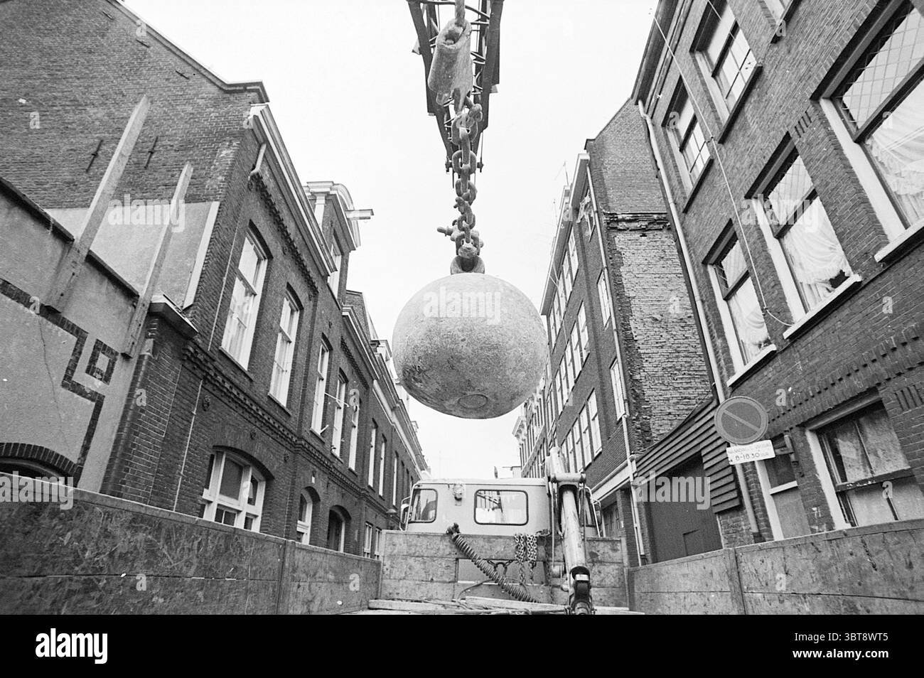 Report Islands Amsterdam Report Amsterdam Grote Bickersstraat pays-Bas, Whizgle News, Dutch Desk, pays-Bas, 1950 - 2000 on 15-06-1972. L'image inclut ces rubriques. La scène est dominée par un imposant objet sphérique soulevé par une grue, suspendu en plein air au-dessus d'une étroite ruelle urbaine. La sphère apparaît lourde, peut-être un matériau de construction ou une installation artistique, et elle contraste fortement avec l'architecture environnante. De chaque côté, les bâtiments en briques reflètent un style urbain classique, caractérisé par leurs rangées ordonnées de fenêtres et leurs nuances variées de bro Banque D'Images