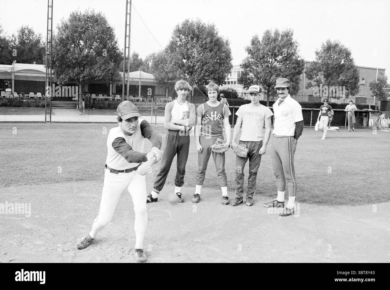 H 'ns Augustinus entraîneur de baseball Baseball Kinheim, Whizgle News, Dutch Desk, pays-Bas, 1950 - 2000 le 05-07-1979. Voici les rubriques de l'image. La scène capture un cadre de baseball extérieur animé, probablement pendant une journée chaude et ensoleillée. Au premier plan, un joueur en uniforme de baseball est capturé à mi-swing, mettant en valeur une pose dynamique qui transmet le mouvement et la concentration. Il porte une casquette, et ses vêtements sont brillants, contrastant avec les tons plus discrets des joueurs environnants. Autour de lui, quatre individus supplémentaires se tiennent décontractés, mettant en valeur un mélange de vêtements athlétiques qui varie en couleurs f Banque D'Images