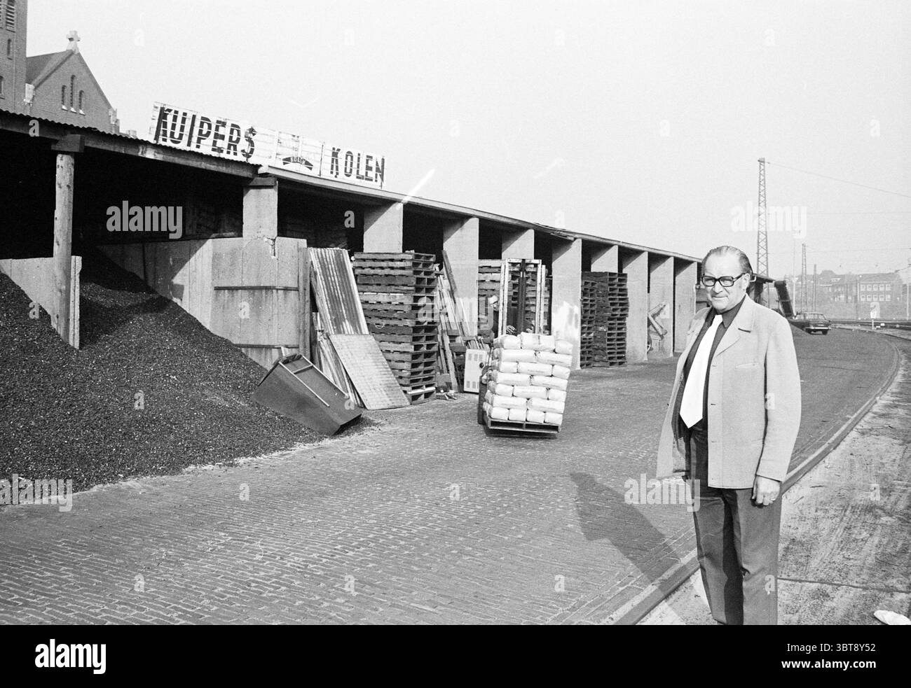 Rapport sur le commerce du charbon de Kuiper, Whizgle News, Dutch Desk, pays-Bas, 1950 - 2000. Ces rubriques apparaissent dans l'image. La scène représente un homme debout au premier plan sur un fond industriel. Il porte une veste de costume de couleur claire sur une chemise plus foncée, complétée par des lunettes qui ajoutent une touche intellectuelle. Son expression semble contemplative alors qu'il regarde légèrement sur le côté. Derrière lui, une grande structure couverte abrite des piles de palettes et de sacs en bois, faisant allusion à une zone de chargement très fréquentée. Les sacs sont soigneusement empilés et semblent contenir un matériau granulaire, peut-être du charbon, qui est Banque D'Images
