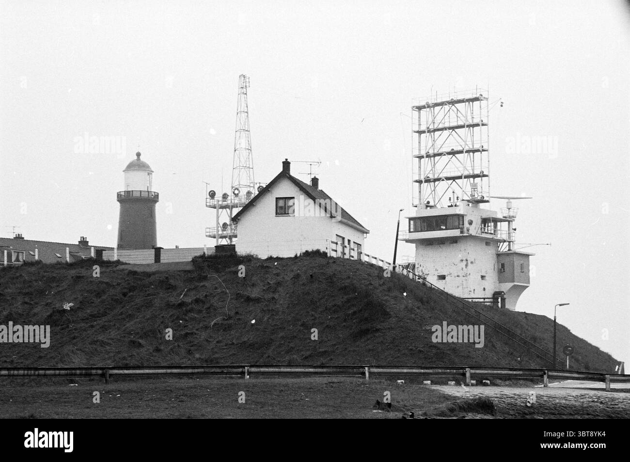 Maison sur Seinpostweg IJmuiden maisons et construction de maisons IJmuiden Seinpostweg pays-Bas, Whizgle News, Dutch Desk, pays-Bas, 1950 - 2000 on 11-03-1981. L'image inclut ces rubriques. La scène présente un paysage côtier caractérisé par un littoral accidenté. À l'avant-garde, une petite maison blanche se trouve sur un tricot herbeux, son architecture simple se fondant dans le cadre naturel. Le bâtiment a une apparence modeste, avec un toit en pente et est entouré d'une végétation clairsemée, renforçant son charme pittoresque. À gauche, un phare traditionnel se dresse haut, avec un rouge classique Banque D'Images