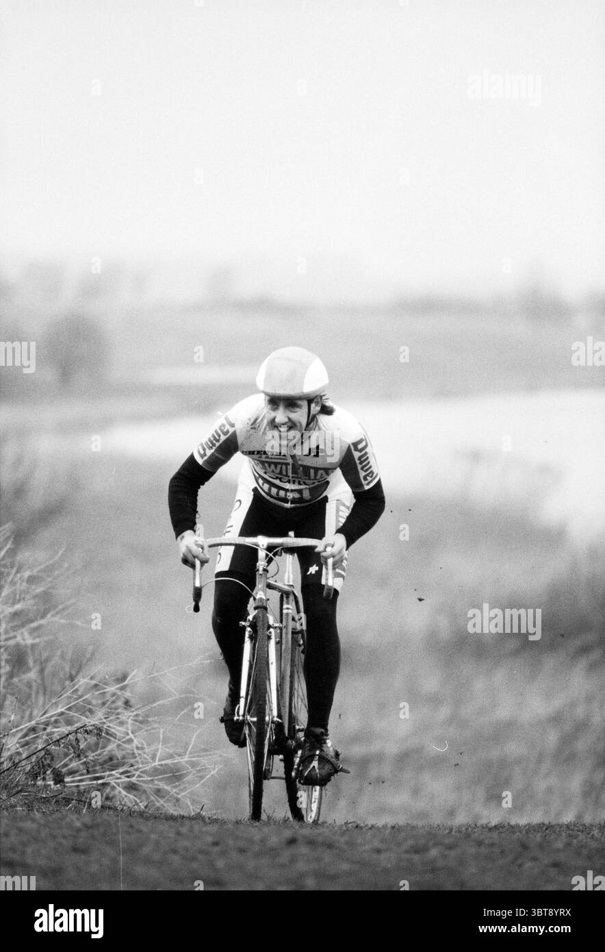 Open cyclo-cross championnat Haarlem pays-Bas, Whizgle News, Dutch Desk, pays-Bas, 1950 - 2000 le 18-01-1992. Ces rubriques sont illustrées dans l'image. Dans la scène, un cycliste est représenté naviguant sur une pente herbeuse. L'individu est vêtu d'une tenue de course vintage ajustée avec des couleurs audacieuses et contrastées, principalement du blanc avec des accents de tons plus sombres. Ils portent un casque profilé, mettant l'accent sur l'accent mis par le sport sur la vitesse. L’expression du cycliste reflète la détermination et l’effort, alors qu’il se penche en avant, pédalant vigoureusement. Le vélo a un design classique, avec des pneus minces et Banque D'Images