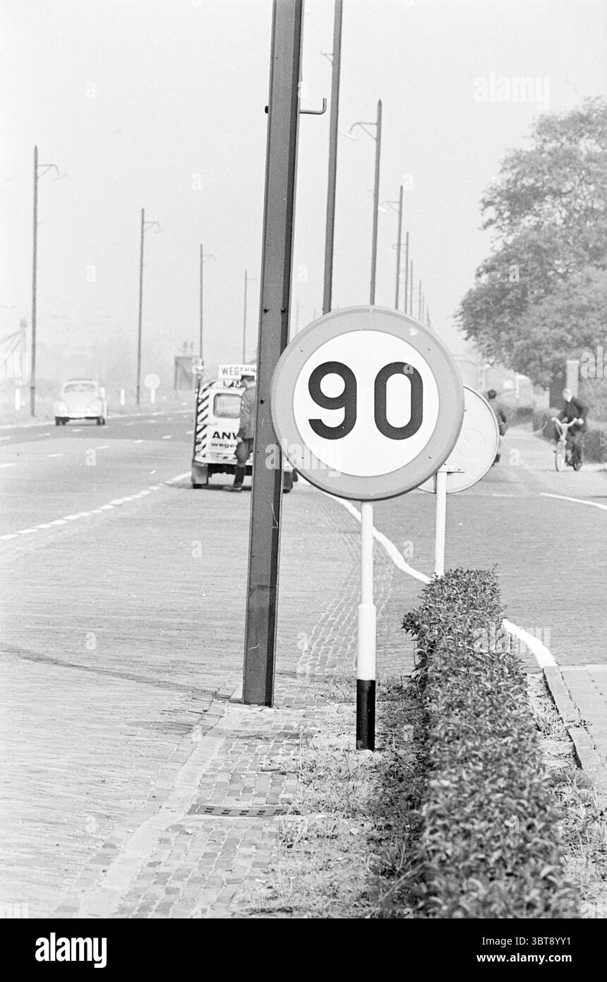 Panneau 90 km Amsterdamse Vaart Traffic Safe Traffic Haarlem Amsterdamsevaart pays-Bas, Whizgle News, Dutch Desk, pays-Bas, 1950 - 2000 on 06-08-1963. L'image inclut ces rubriques. La scène capture un tronçon tranquille de route flanqué d'arbustes bas et d'herbe, suggérant une zone rurale ou moins développée. Au premier plan se trouve un panneau circulaire affichant le chiffre « 90 », placé bien en évidence sur un poteau. L'arrière-plan blanc gras de l'enseigne contraste avec le grand chiffre noir, attirant l'attention immédiatement. En arrière-plan, un seul véhicule, éventuellement un camion, est stationné à côté Banque D'Images